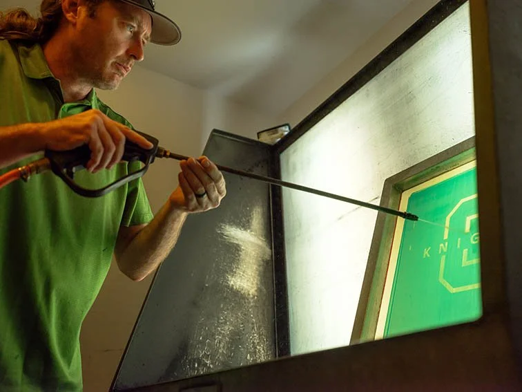 A man in a green shirt using a spray gun to clean a green screen.