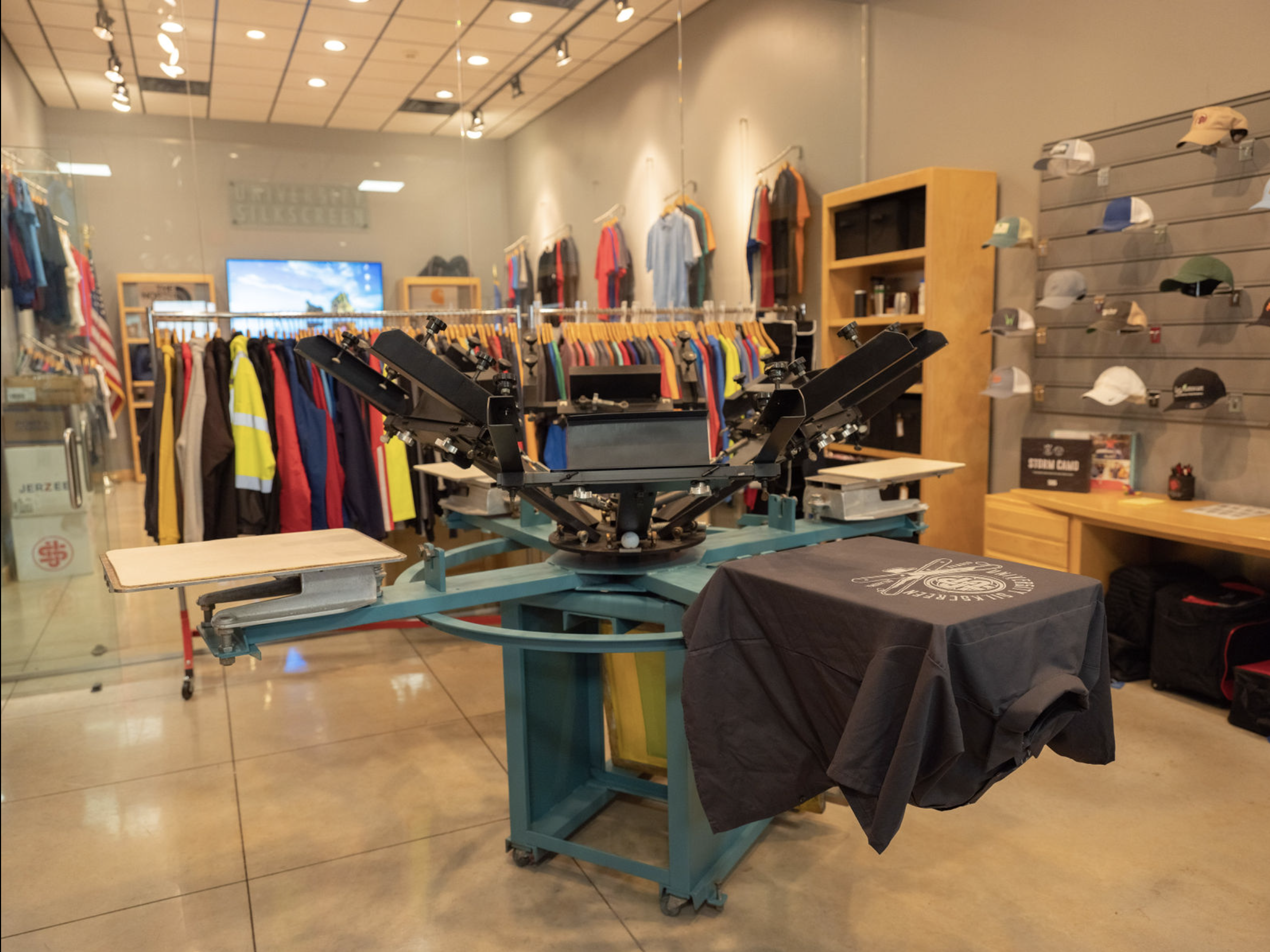 Screen-printing shop setup showcasing apparel, hats, and a press in action.