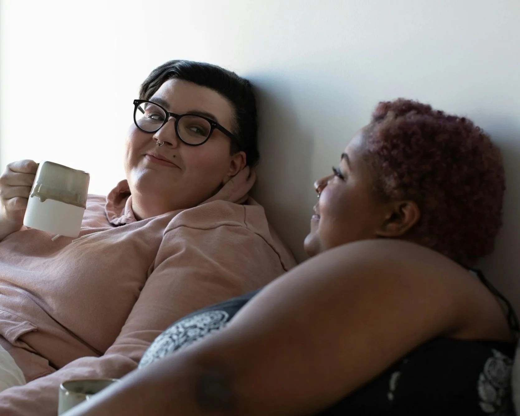 Two women sitting close together, one holding a mug, smiling and looking at each other in a cozy indoor setting.