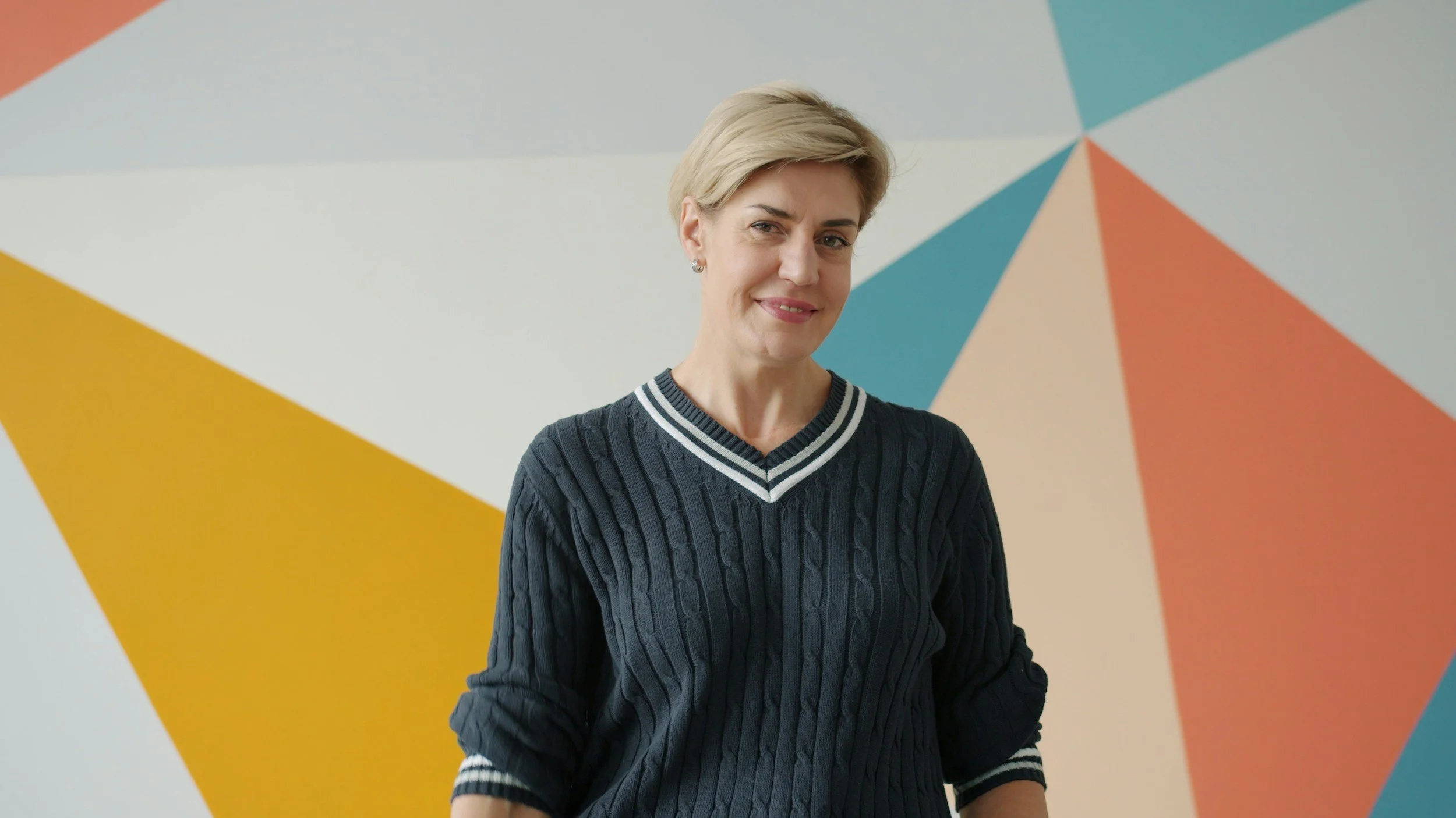 A woman with short blonde hair wearing a dark cable-knit sweater with white and black striped trim, standing in front of a colorful geometric mural.