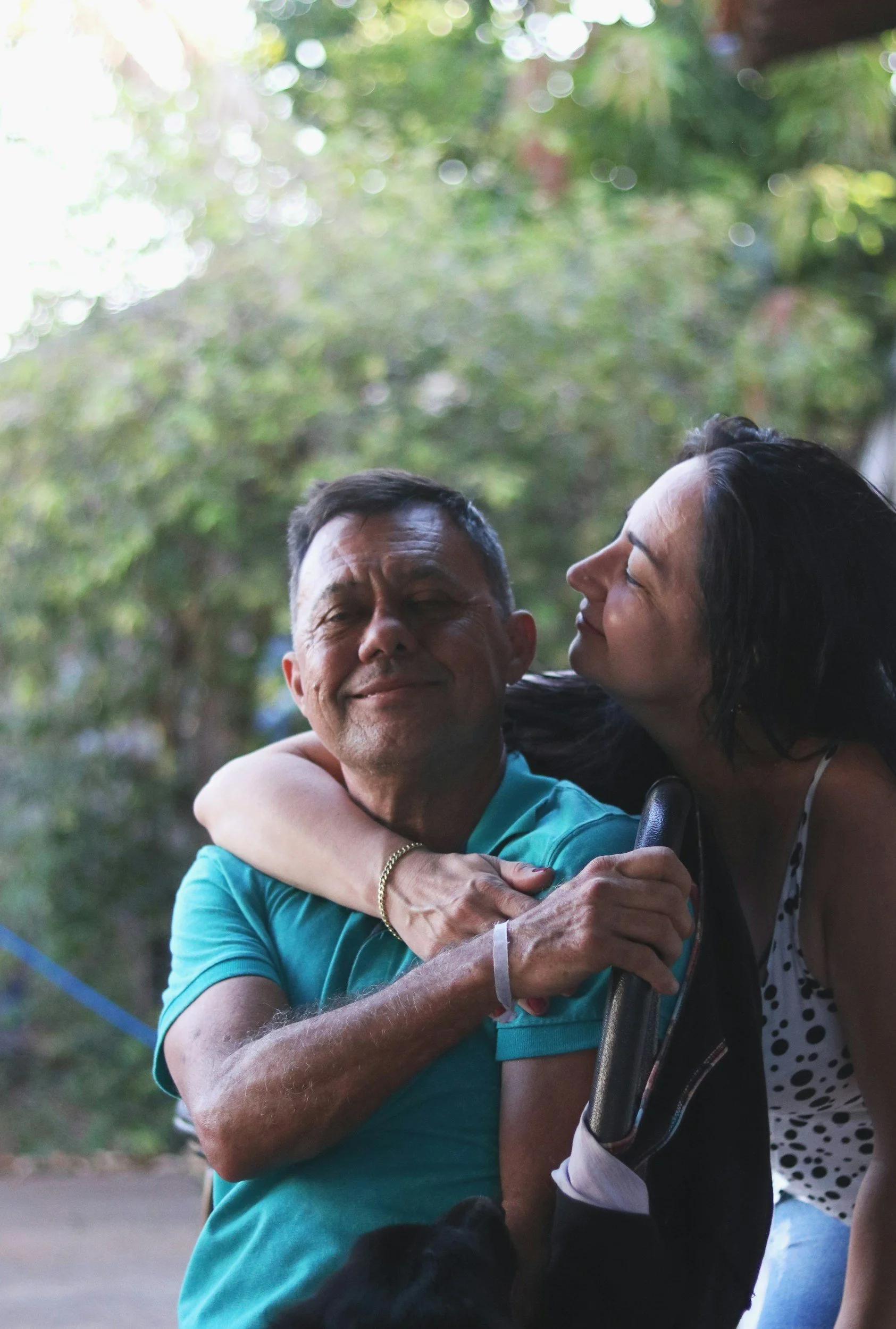 A woman hugging a man who appears to be in a wheelchair, both smiling, in a green outdoor setting.