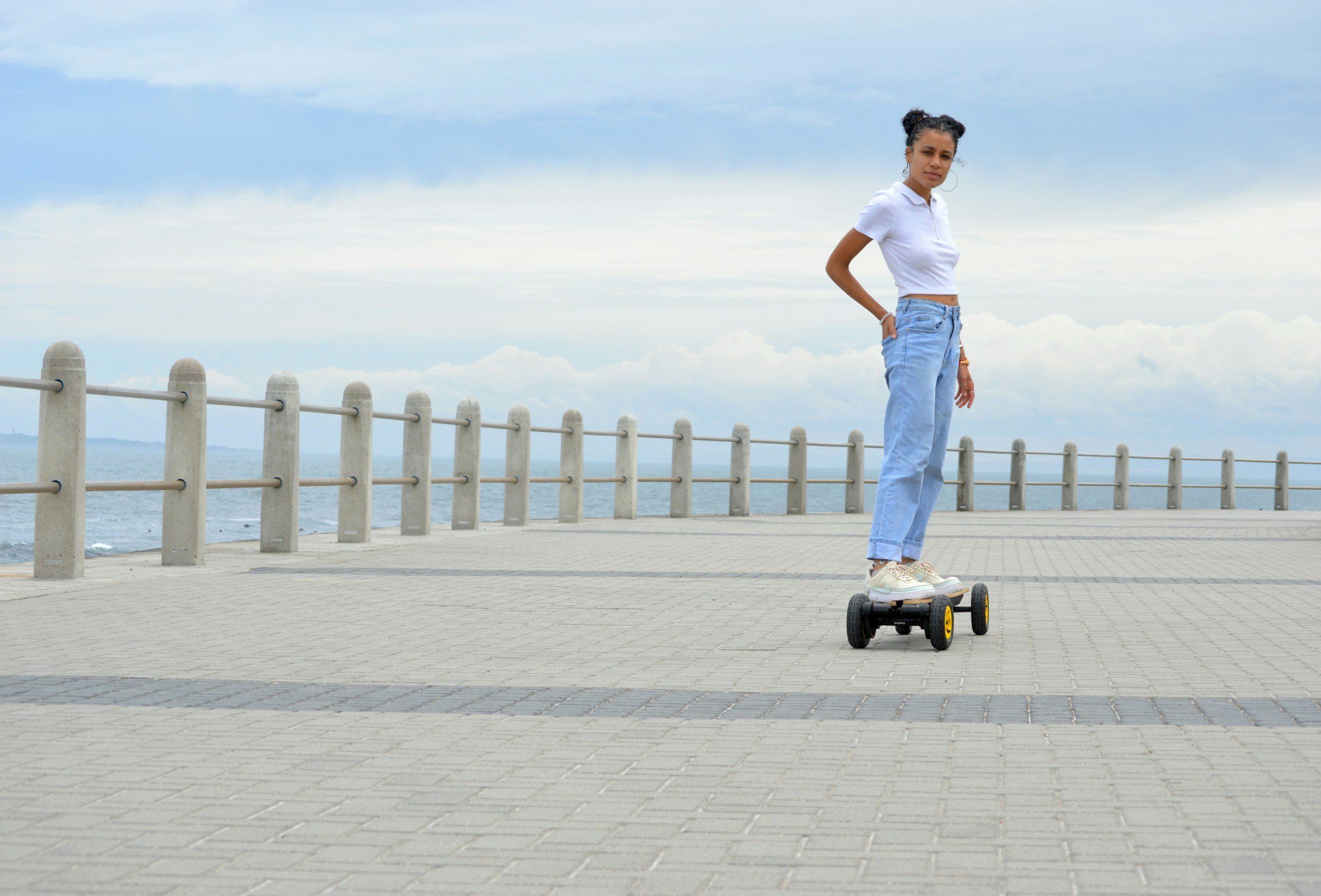 Young woman standing on a skateboard on a paved promenade by the ocean with a cloudy sky overhead.
