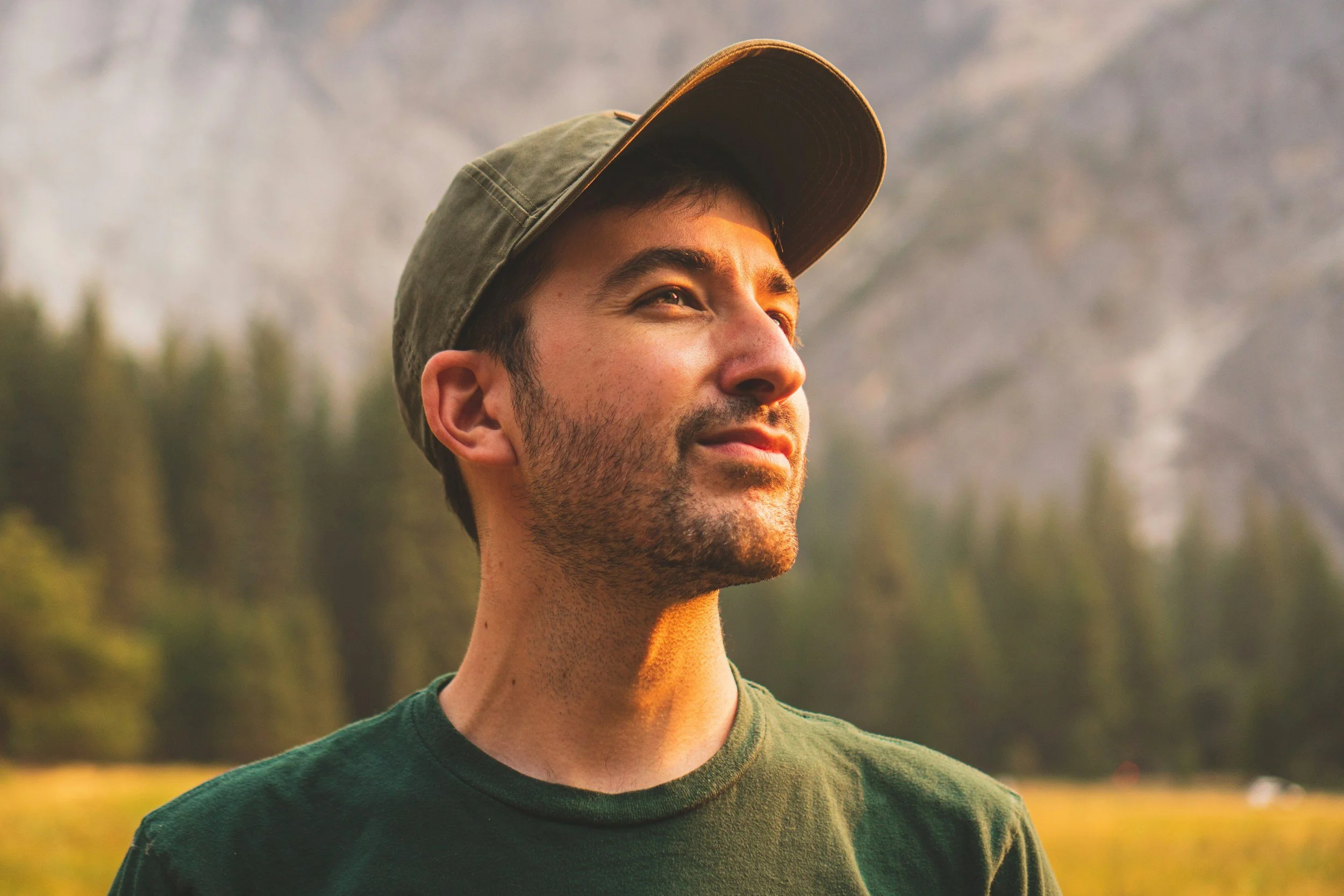 A man wearing a green t-shirt and a brown cap, looking off into the distance with a serene expression, outdoors with a background of trees and mountains.