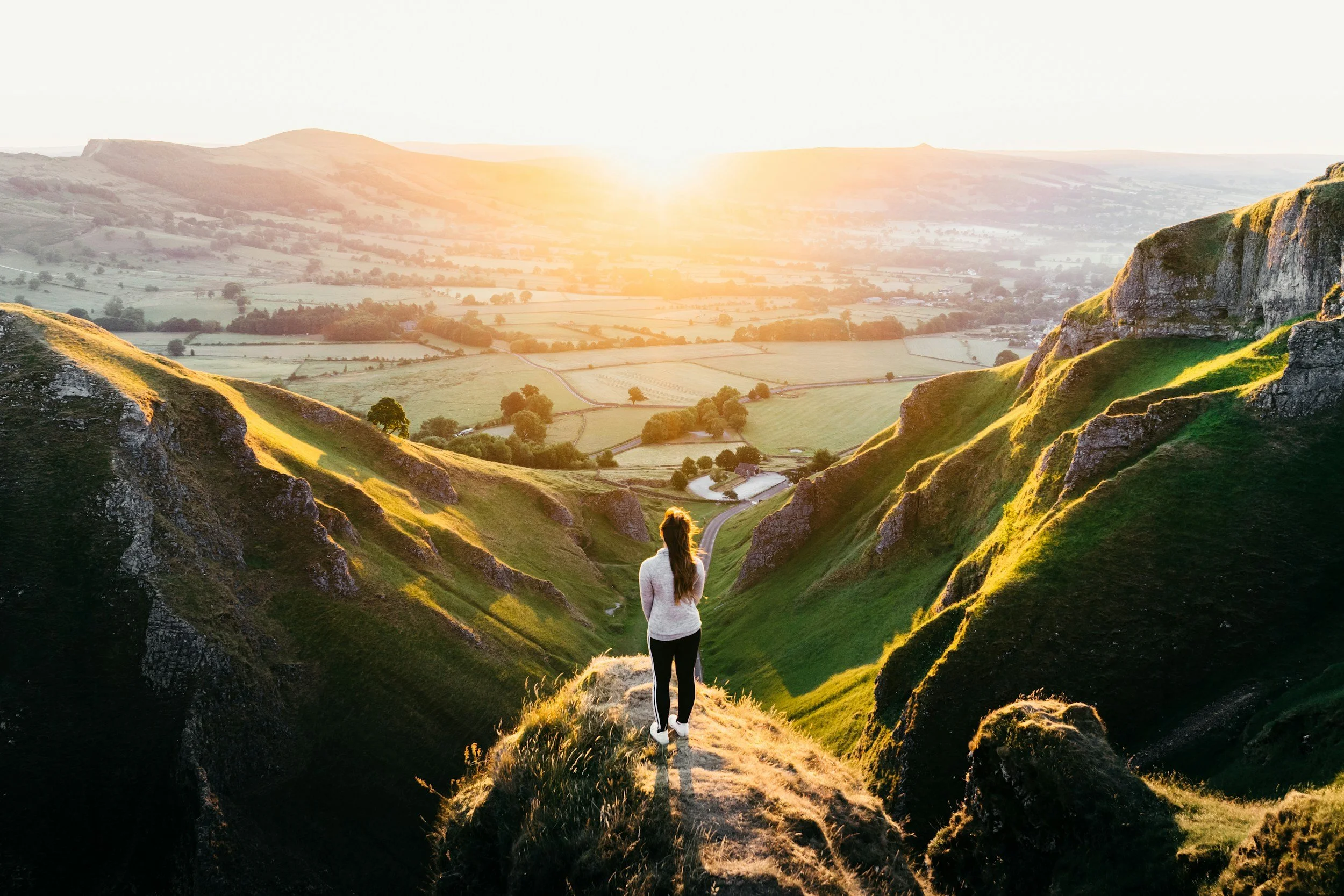 A woman stands on a narrow trail between green hills, overlooking a picturesque valley at sunset with rolling fields and a few trees.