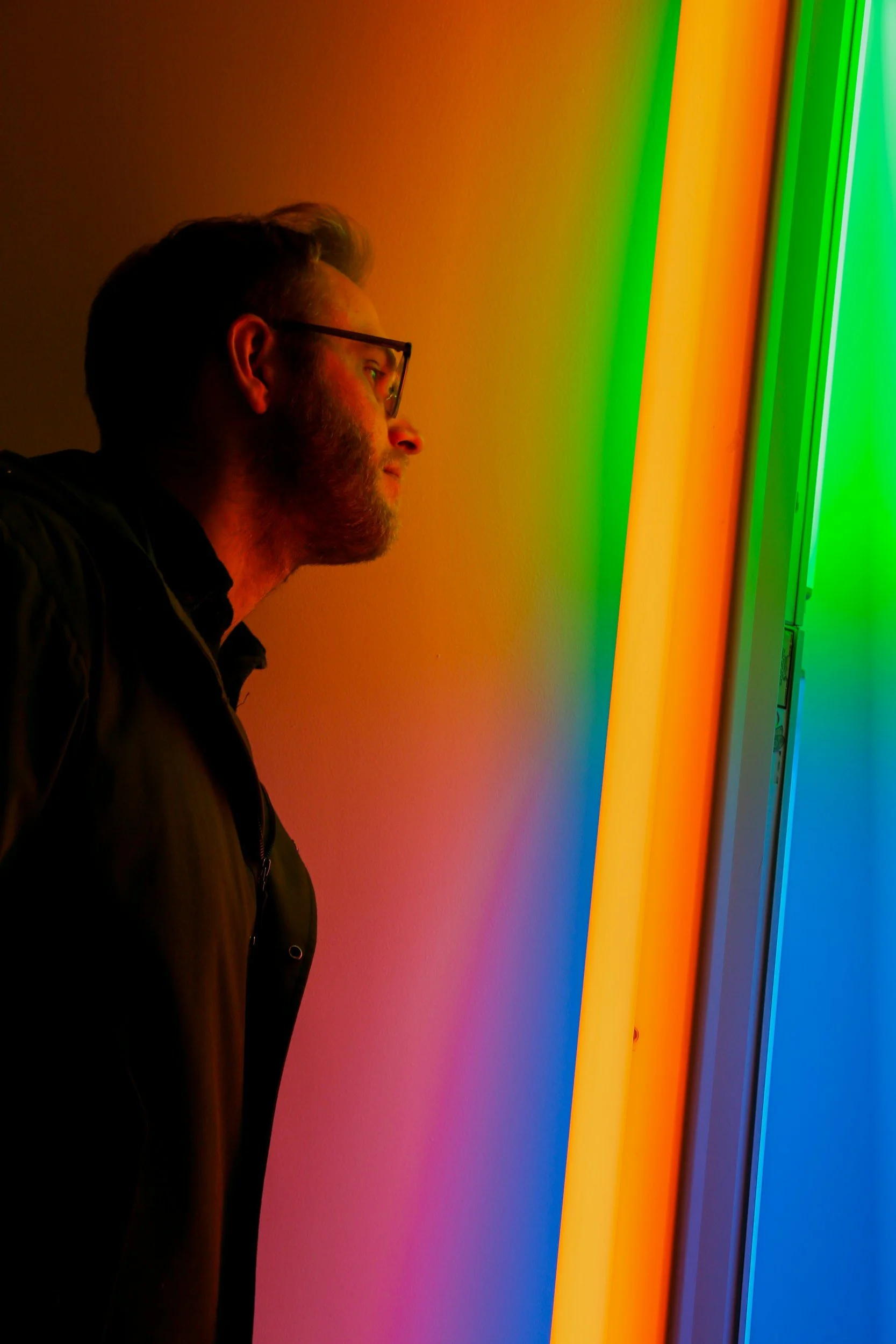 A man with glasses and a beard is looking at a colorful rainbow light display.