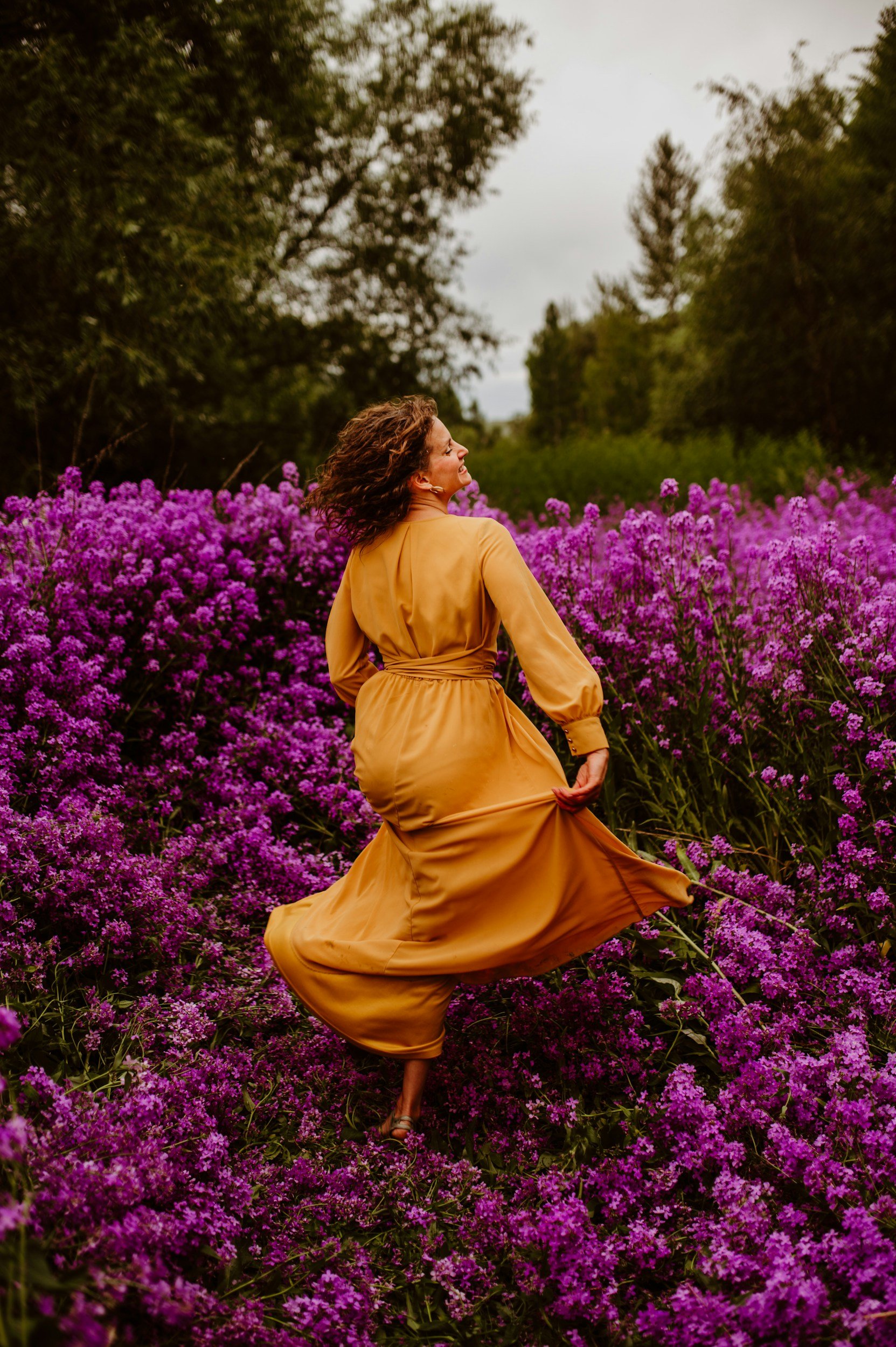 A woman in a light brown dress walking through a field of purple flowers, with trees in the background on an overcast day.