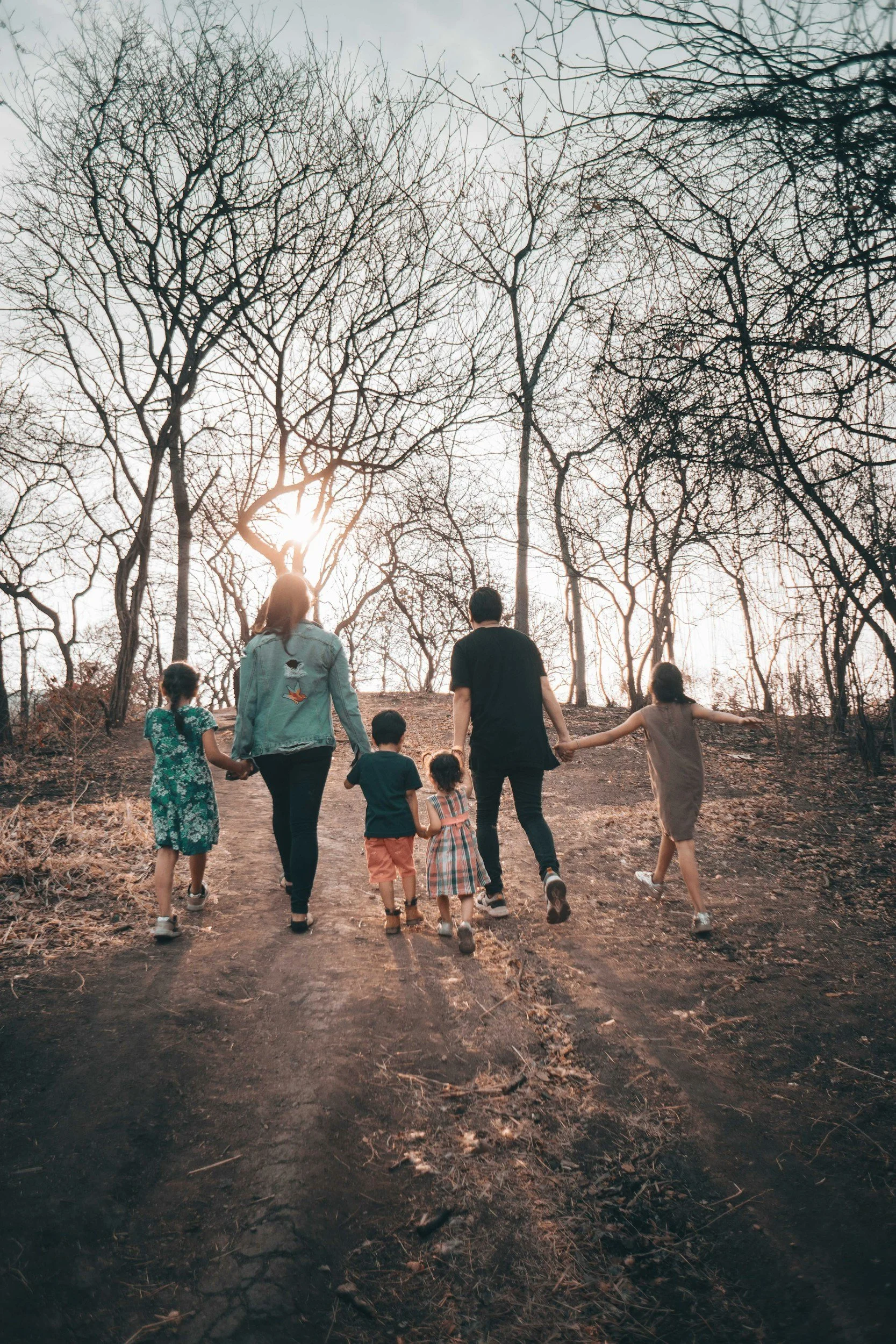 Family walking hand in hand on a dirt trail in a leafless forest during sunset.