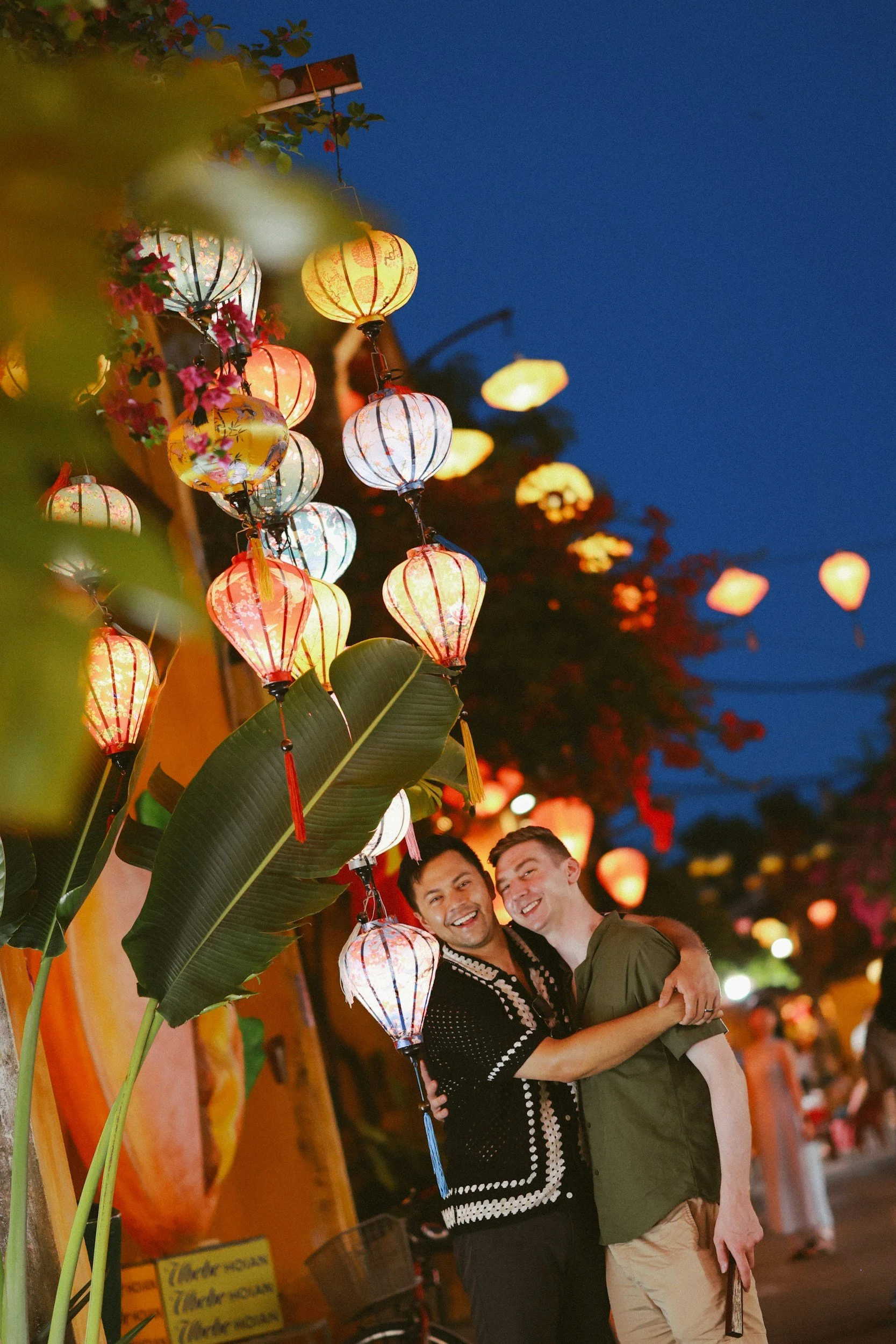 Two men smiling and hugging under colorful paper lanterns hanging at night, with a vibrant street scene in the background.