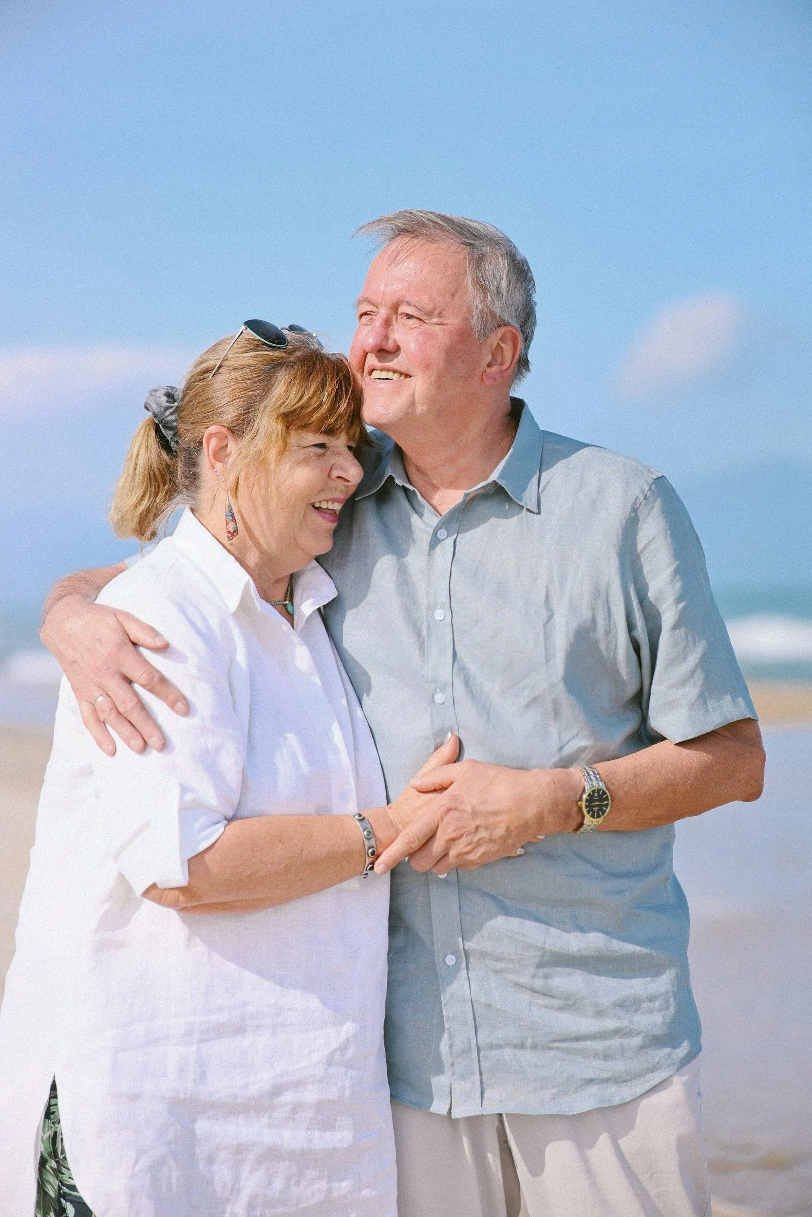 An elderly couple hugging and smiling on a sunny beach with blue skies.