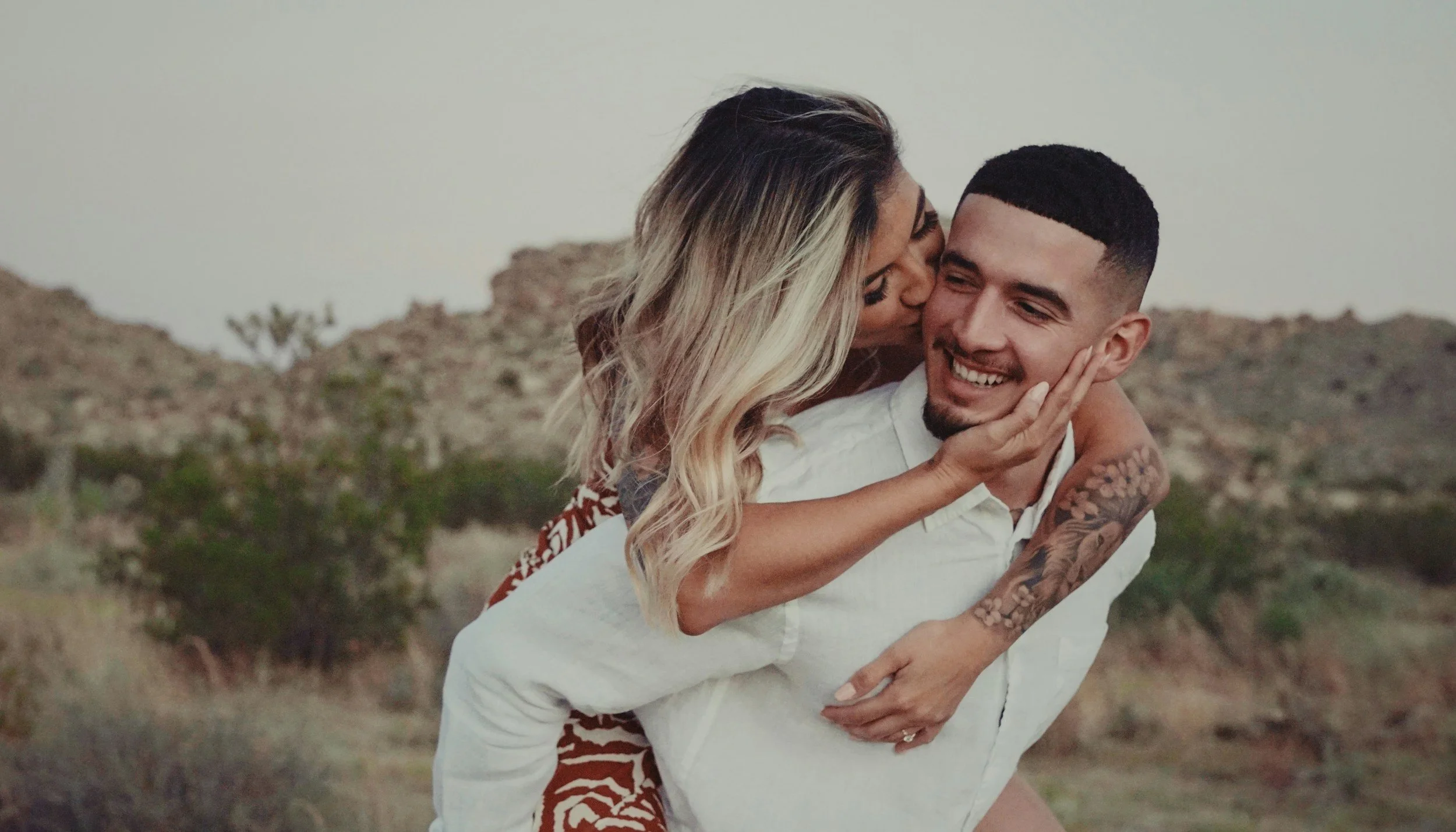 A woman with blonde hair and a tattooed arm kisses a smiling man on the cheek. The man has short dark hair, a beard, and is wearing a white shirt. They are outdoors in a desert-like landscape with hills and sparse vegetation.
