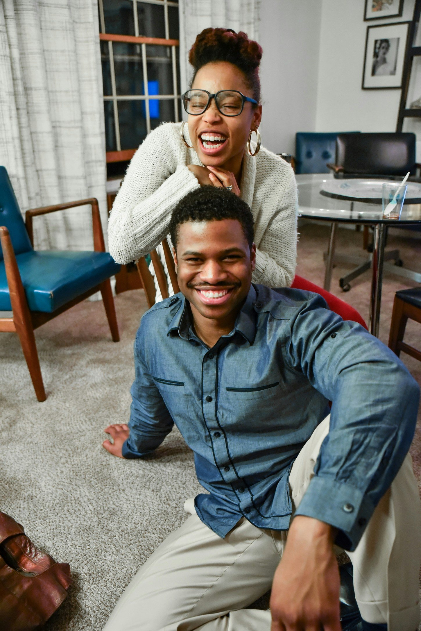 A woman and a man smiling indoors, with the woman leaning on the man's shoulder as he sits on the carpeted floor.