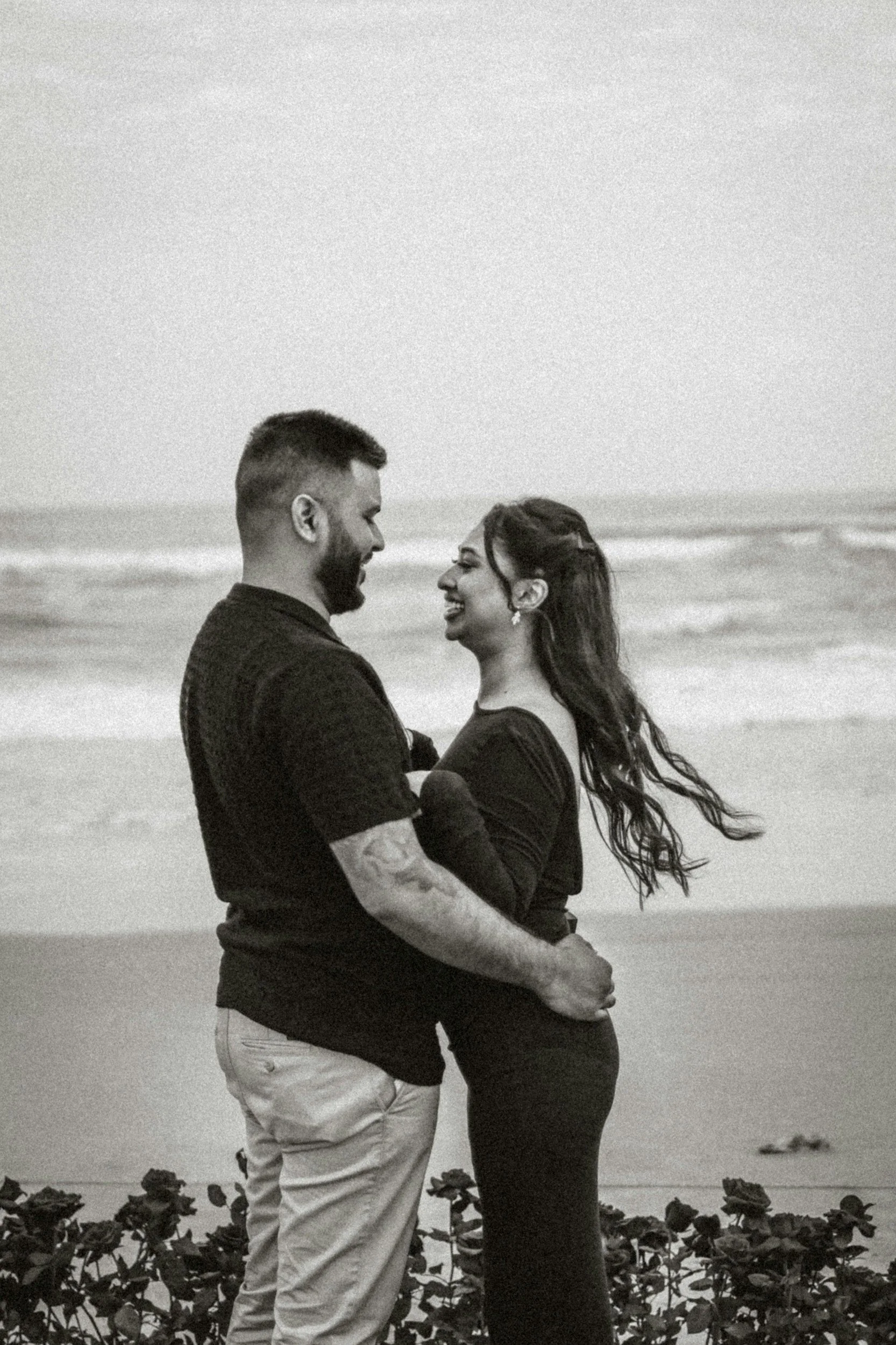 A couple standing close together on a beach, smiling and looking into each other's eyes, with ocean waves in the background.