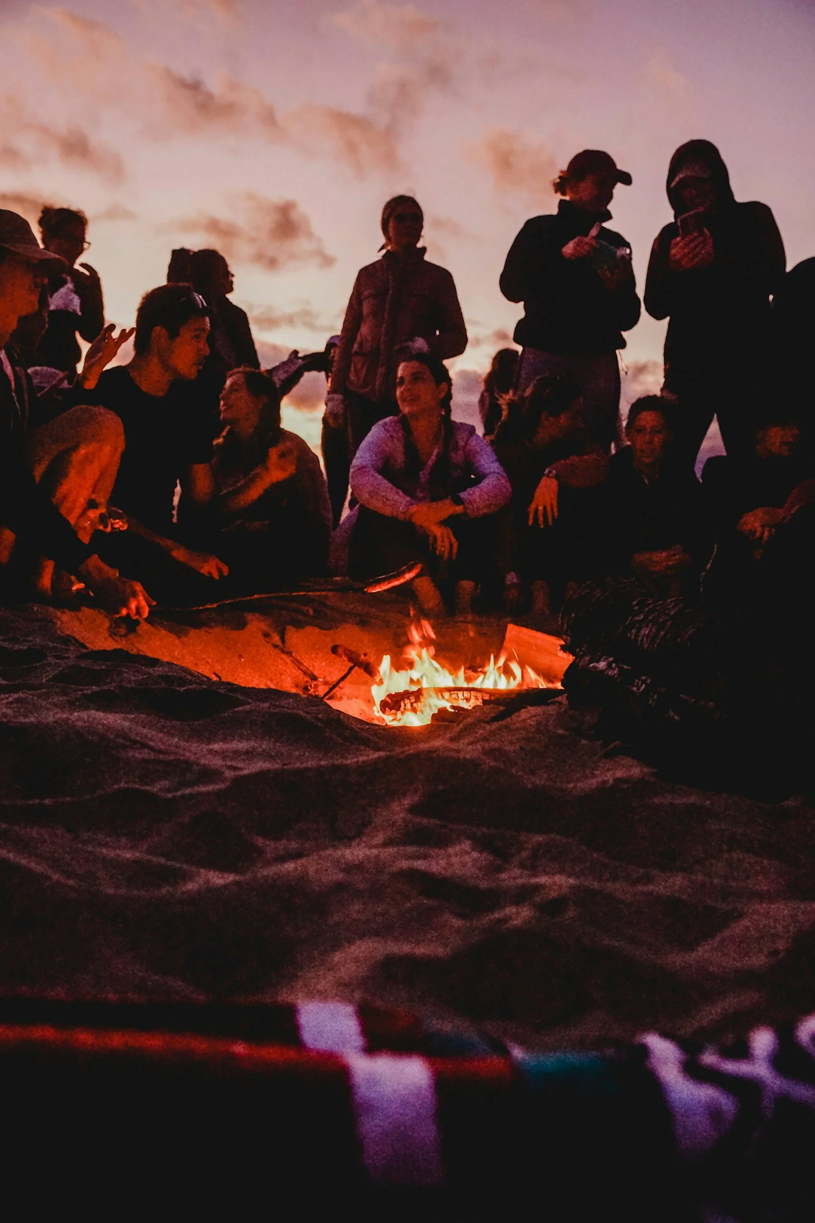 A group of people gathered around a campfire outdoors during sunset, some sitting and some standing, engaging in conversation.