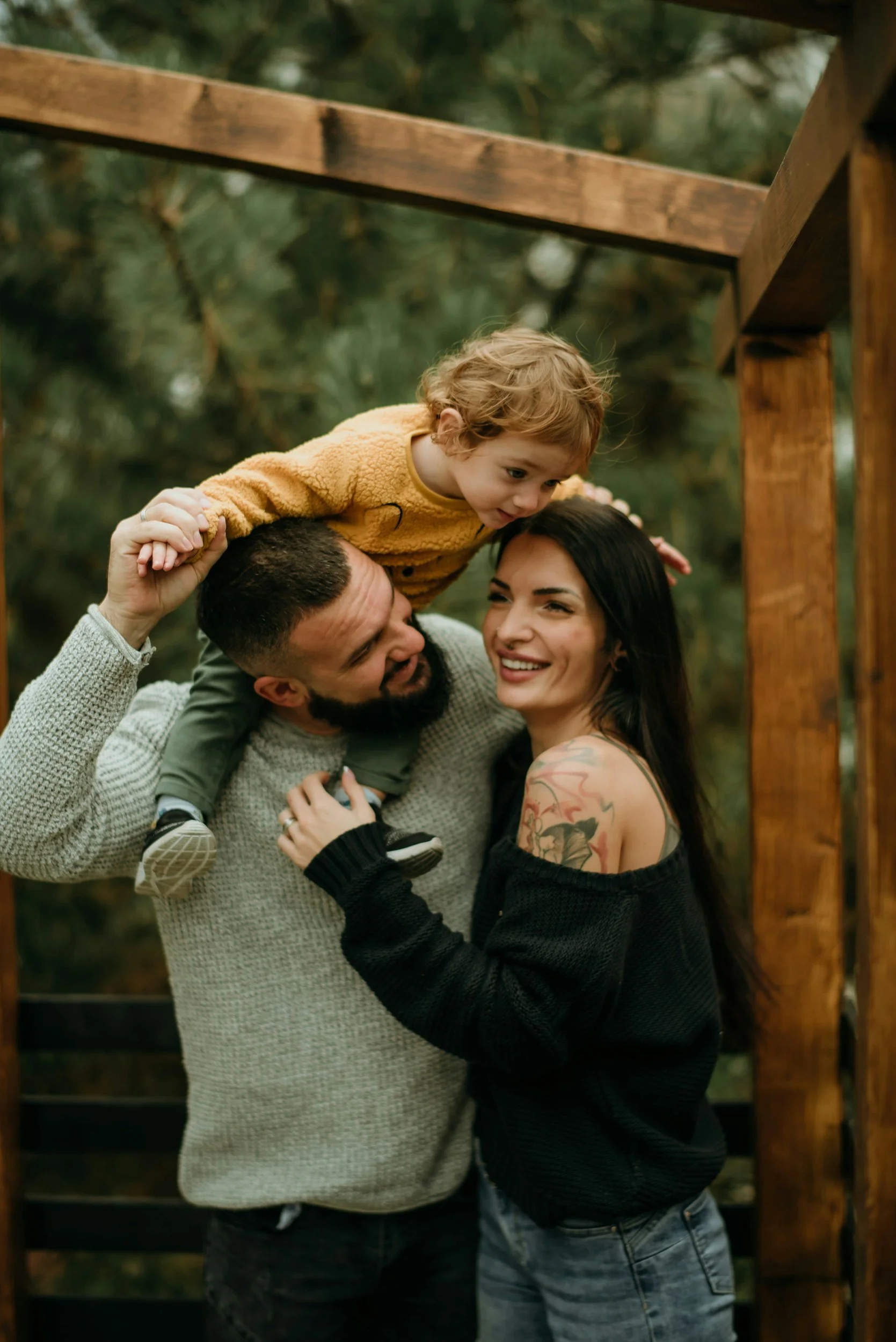 Family of three outdoors under a wooden structure, with the father lifting the young child onto the mother's shoulders, all smiling.