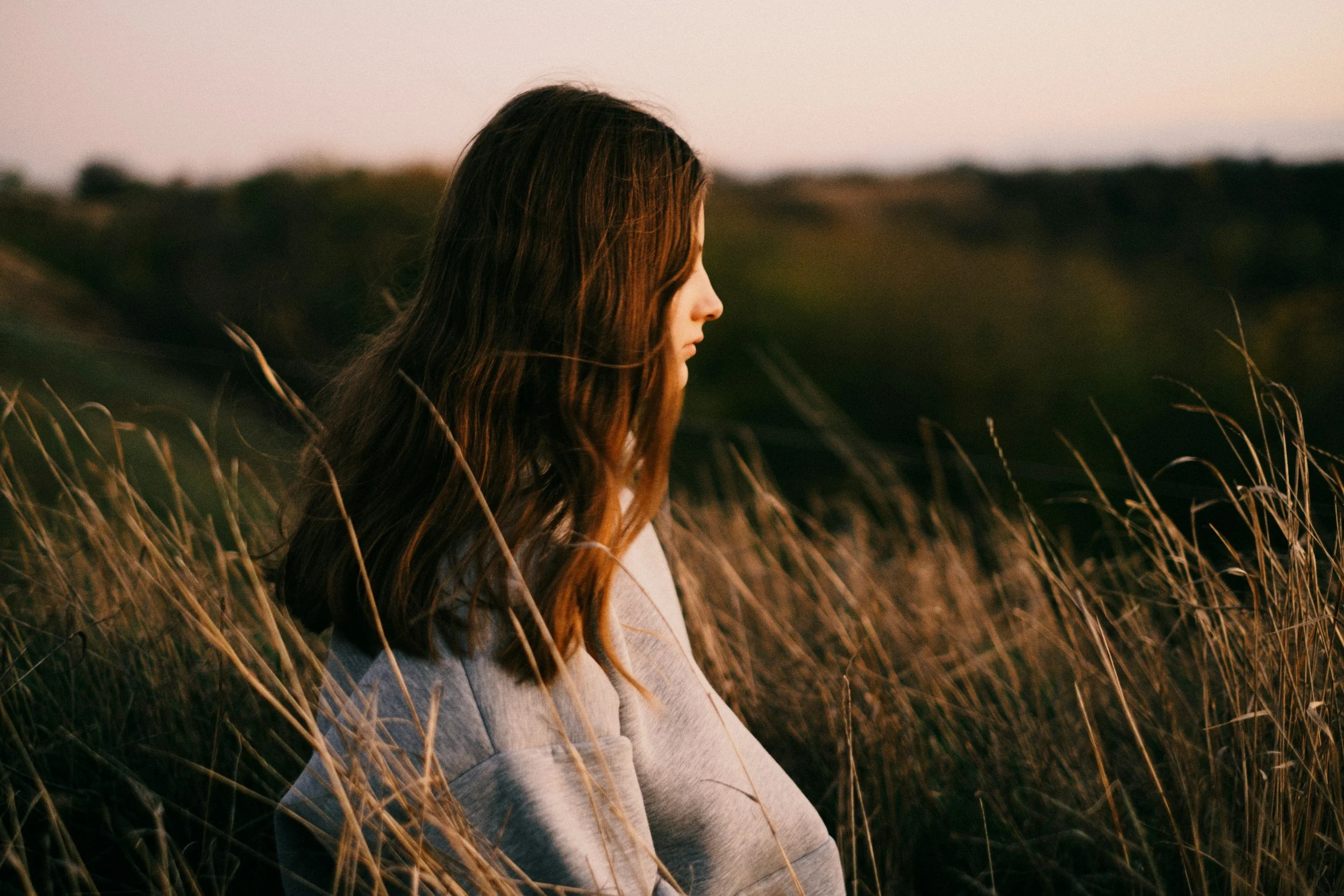 A woman with long brown hair standing in a field of tall, dry grass during sunset, facing to the right in profile.