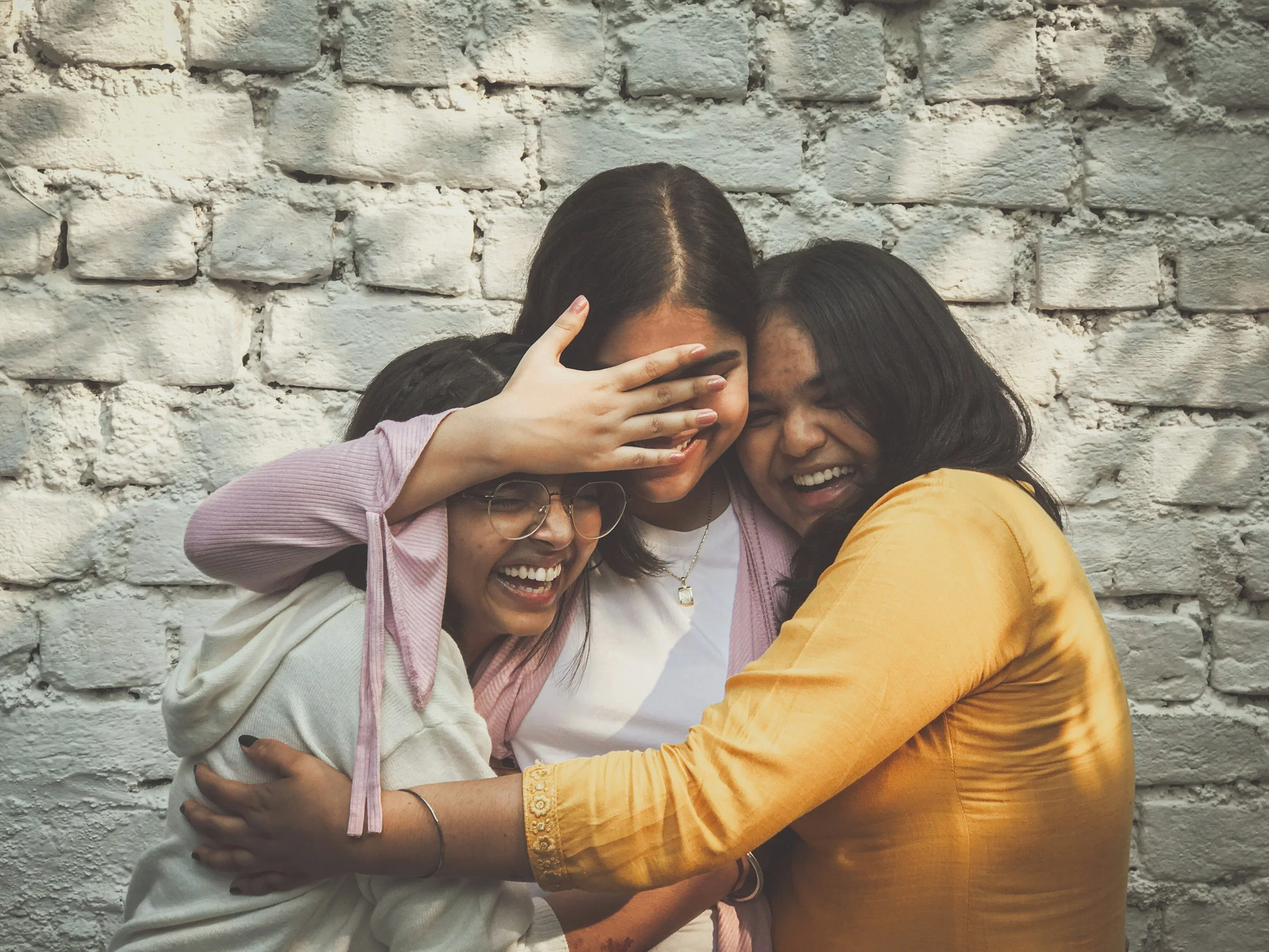 Three women laughing and hugging against a white brick wall.
