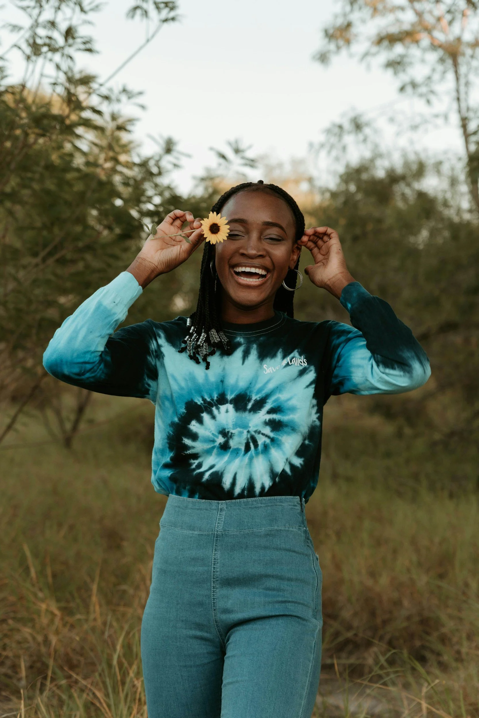 A woman smiling outdoors, holding a sunflower to her temple, wearing a black and blue tie-dye sweatshirt and a high-waisted denim skirt, with trees and sky in the background.