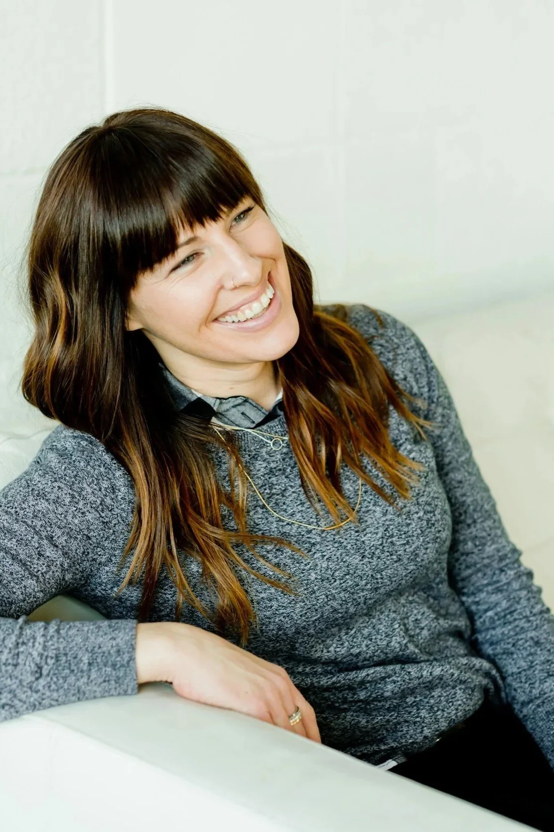 Smiling woman with brown hair sitting on a white couch wearing a gray sweater.