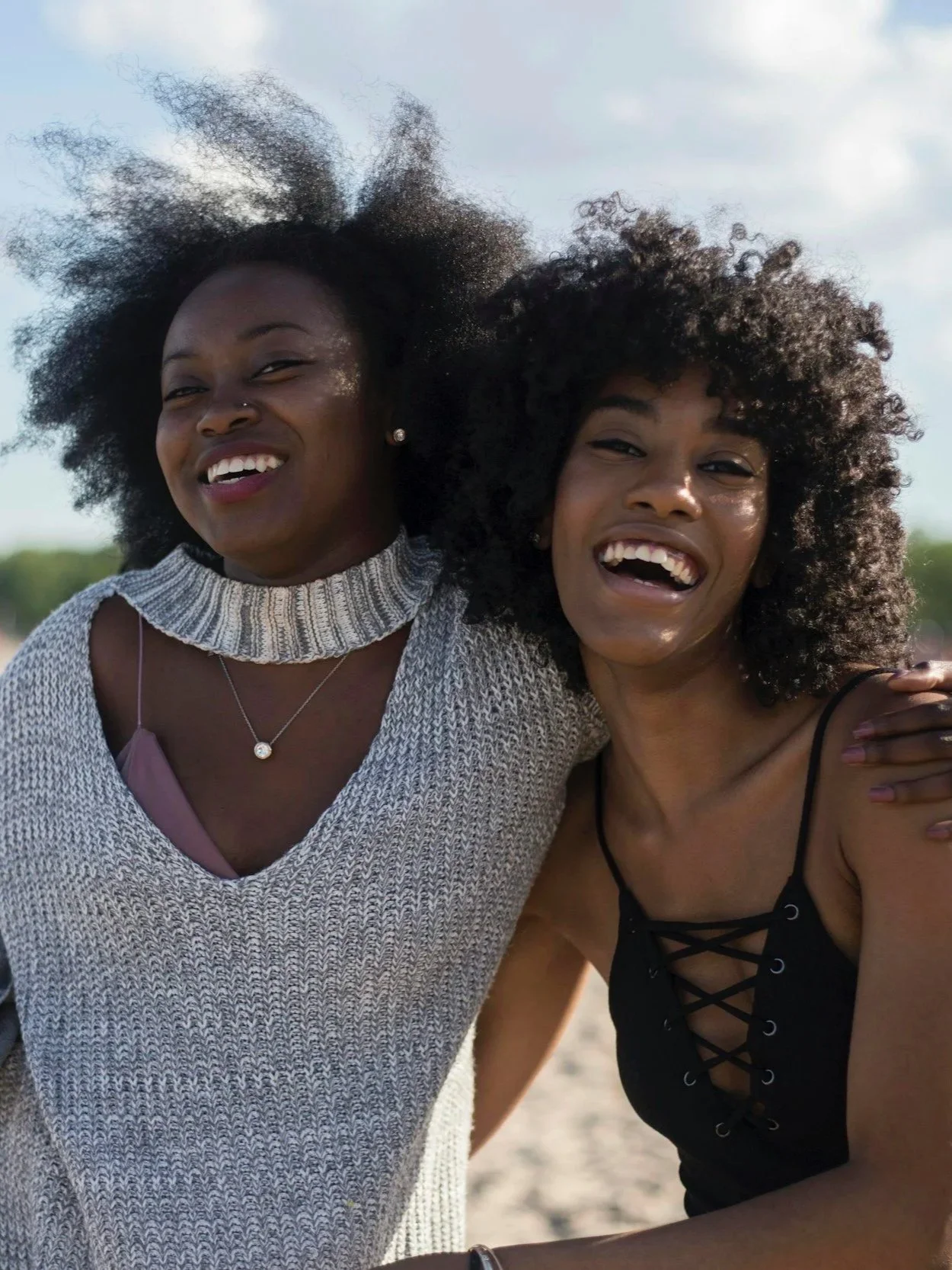 Two women with curly hair smiling and laughing, standing outdoors on a sunny day.