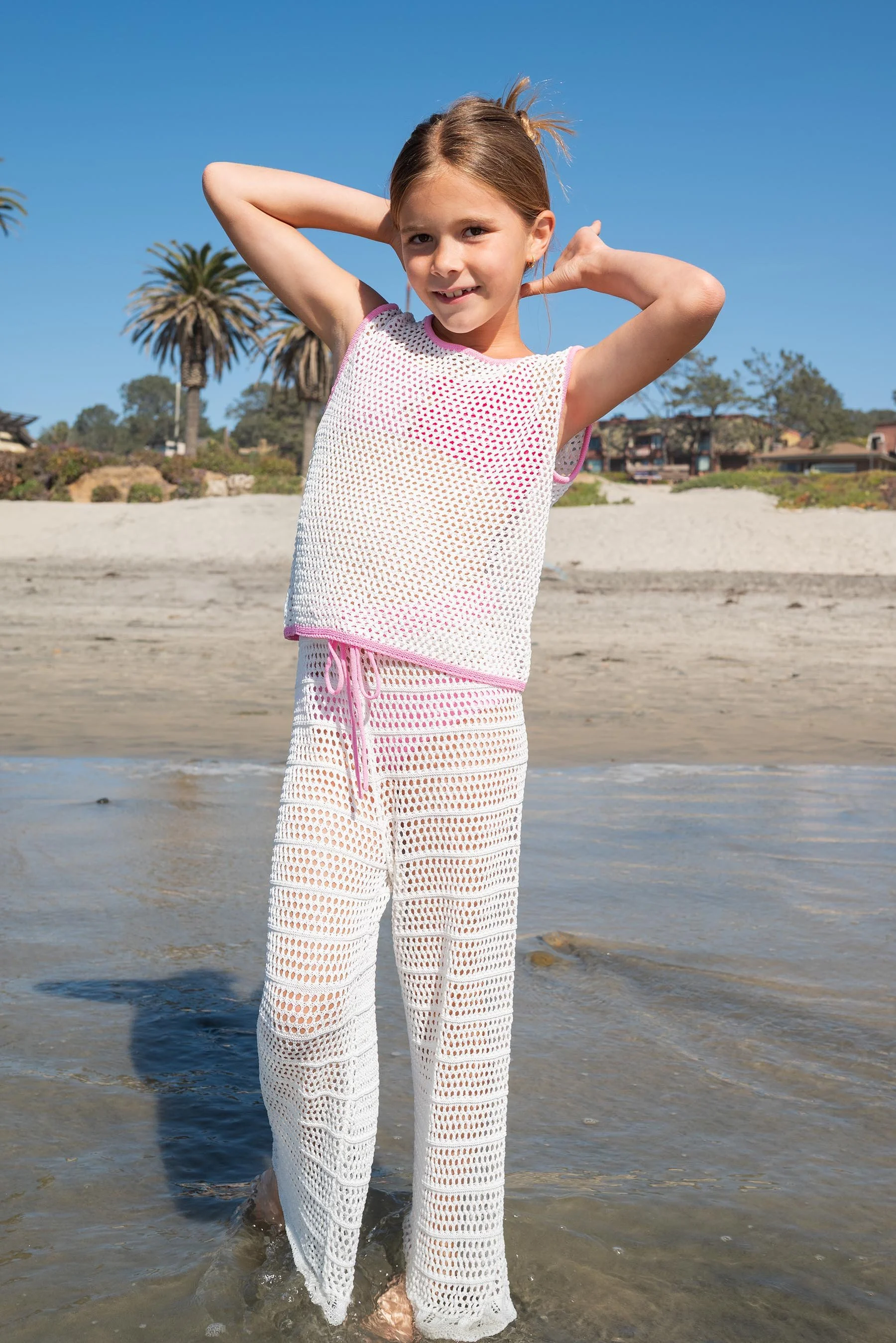 Young girl standing in shallow water at the beach, wearing a white mesh top and matching mesh pants, with palm trees and houses in the background under a clear blue sky.