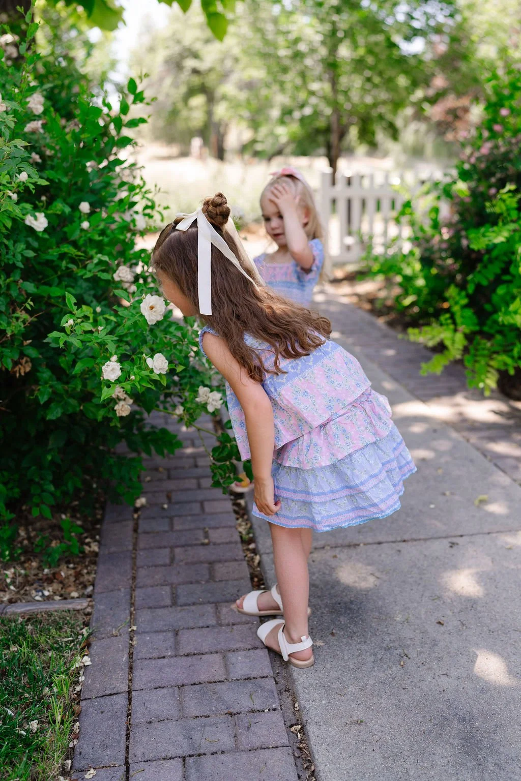 Two young girls in pastel dresses exploring a garden with white blooming flowers, one girl smelling the flowers and the other looking at her head with a hand on her forehead, on a sunny day.