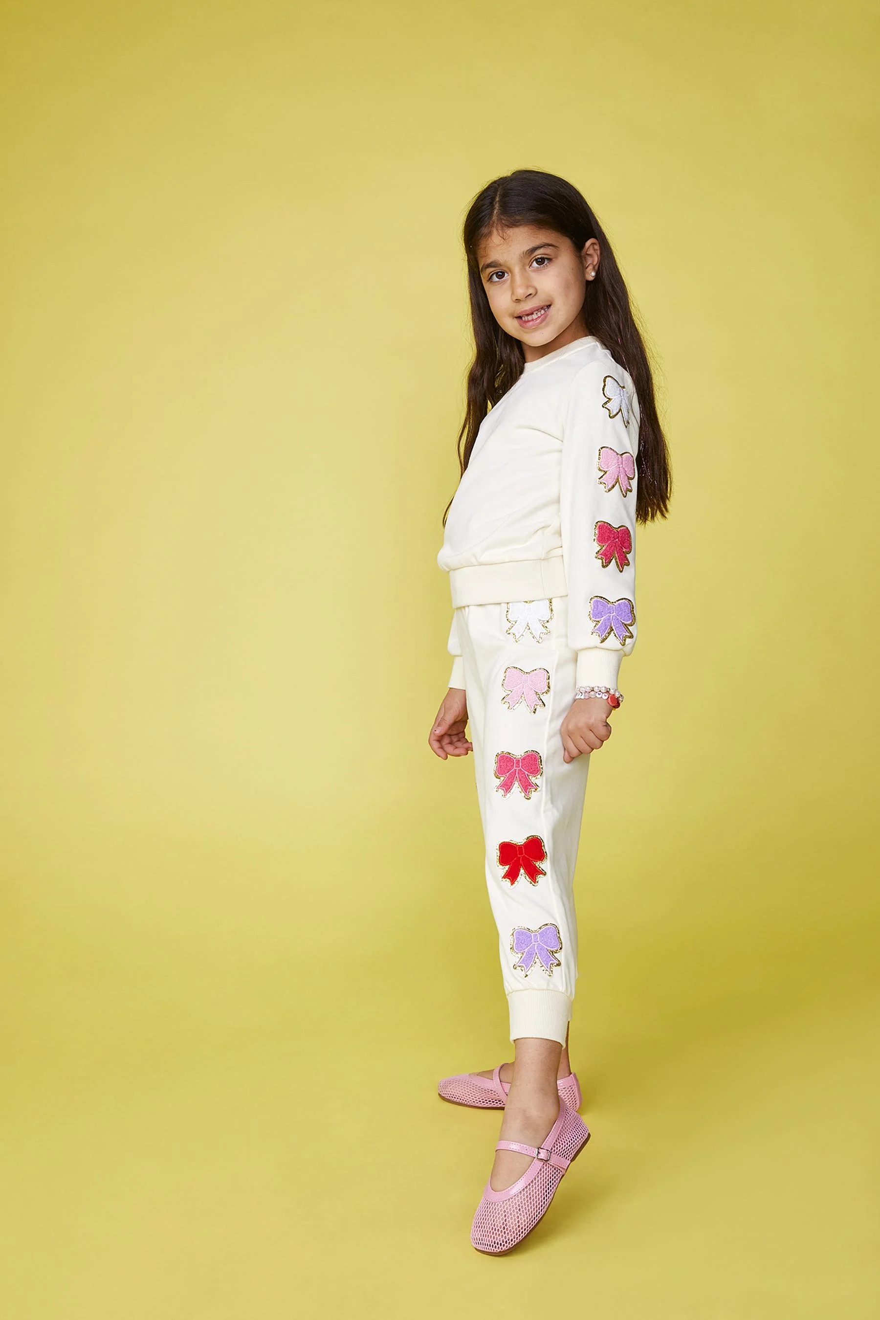 A young girl with long brown hair wearing a white outfit with colorful bow designs on the sleeves and pants, standing against a yellow background, smiling and looking at the camera.