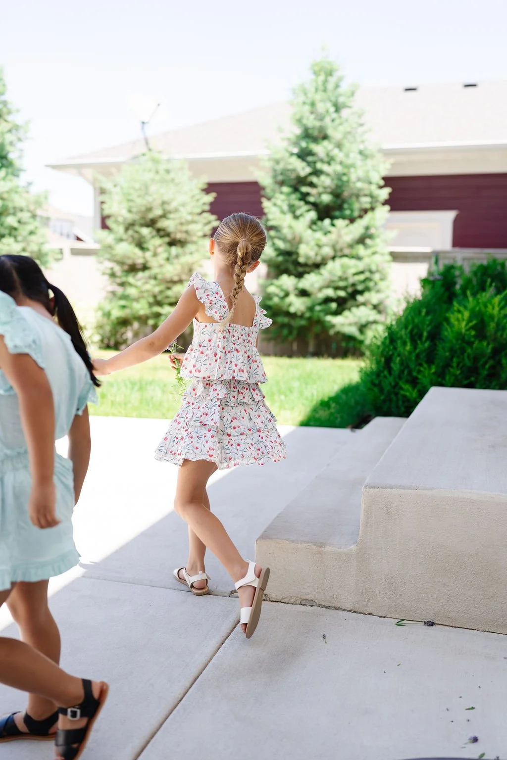 Two young girls in summer dresses playing outside on a sunny day near steps with trees and a house in the background.