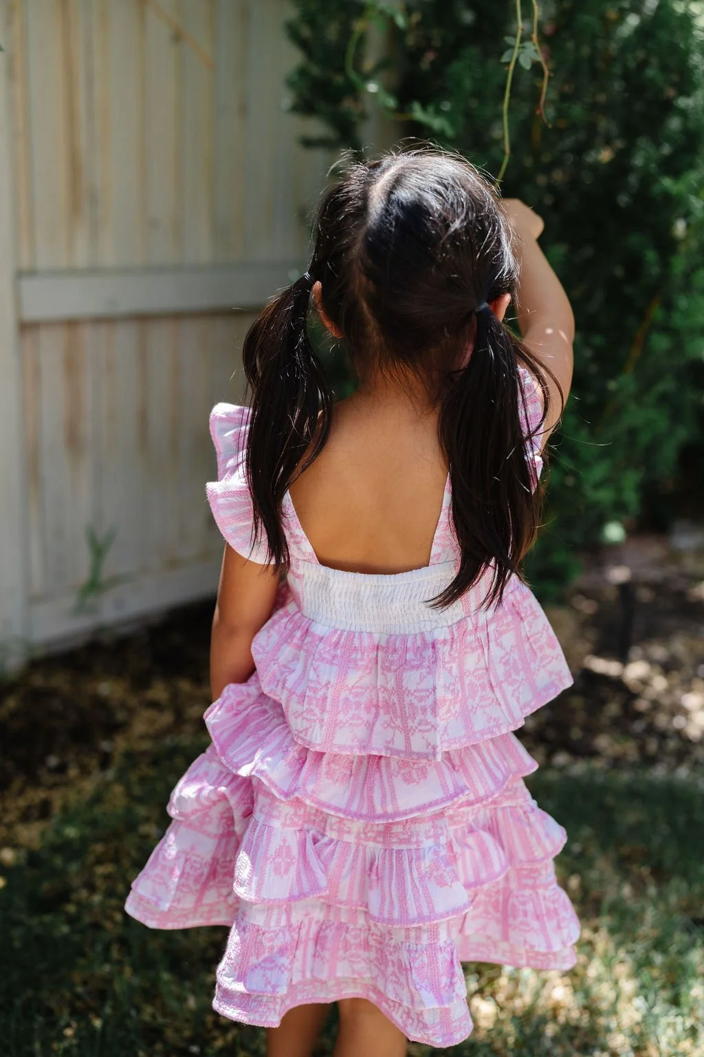 Back view of a young girl with long dark hair in pigtails, wearing a pink and white ruffled dress, outdoors next to a wooden fence and green bushes.