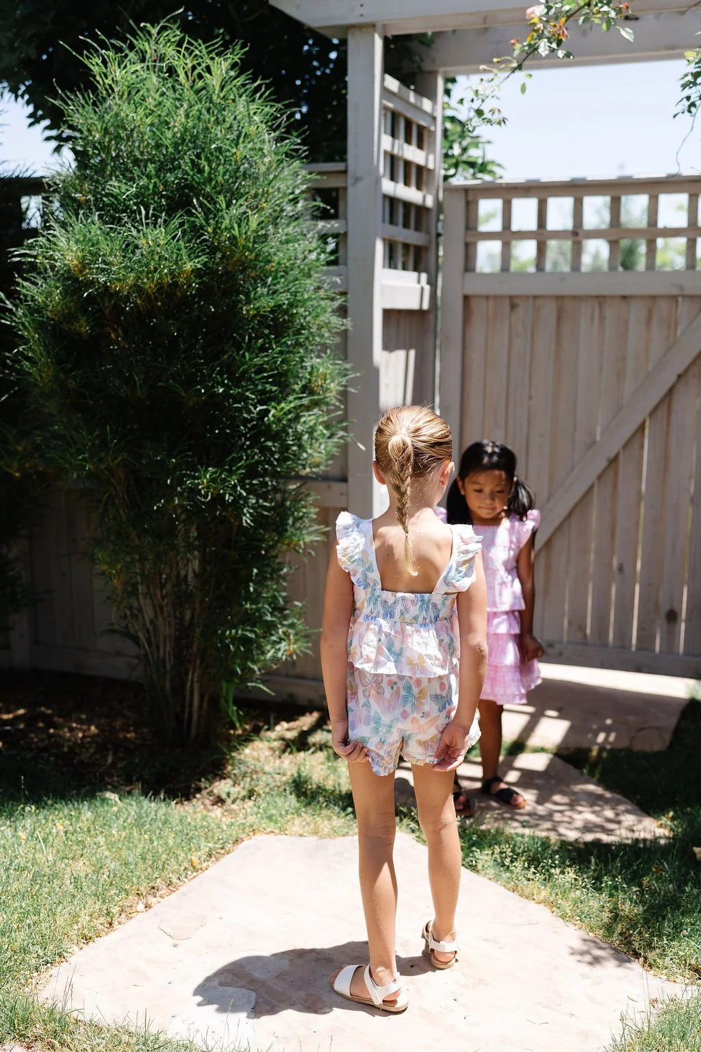 Two young girls in summer clothing standing on a concrete pathway, outside next to a wooden fence, with one girl looking toward the other near the gate.