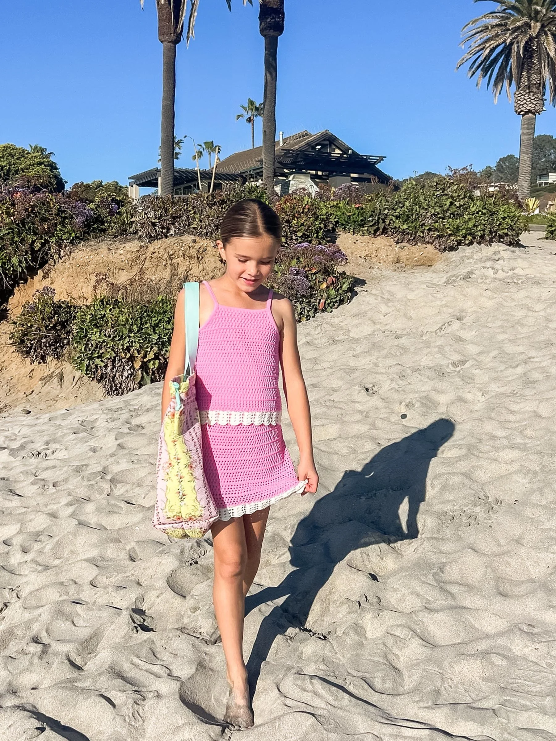Young girl in a pink crochet dress walking on sandy beach with palm trees and a building in the background.