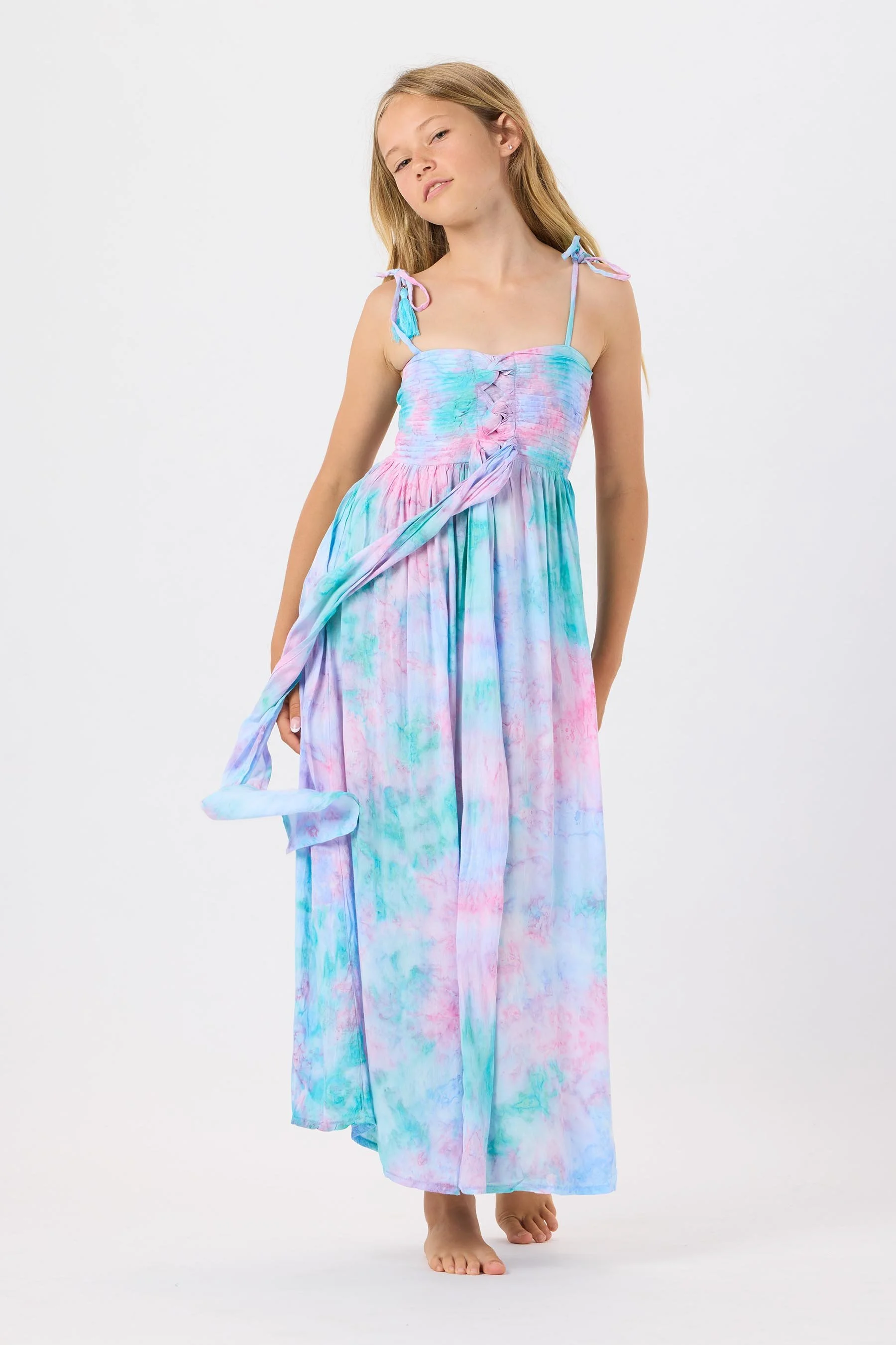 Young girl wearing a pastel tie-dye maxi dress with spaghetti straps and tie shoulder details, standing barefoot against a plain white background.
