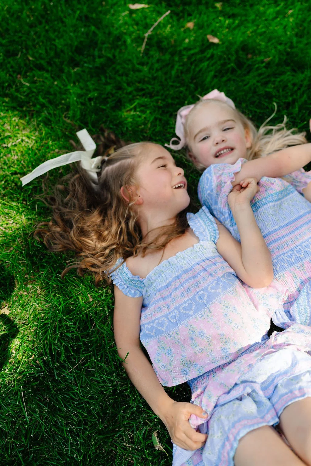 Two young girls lying on green grass, smiling and laughing together in a playful moment.