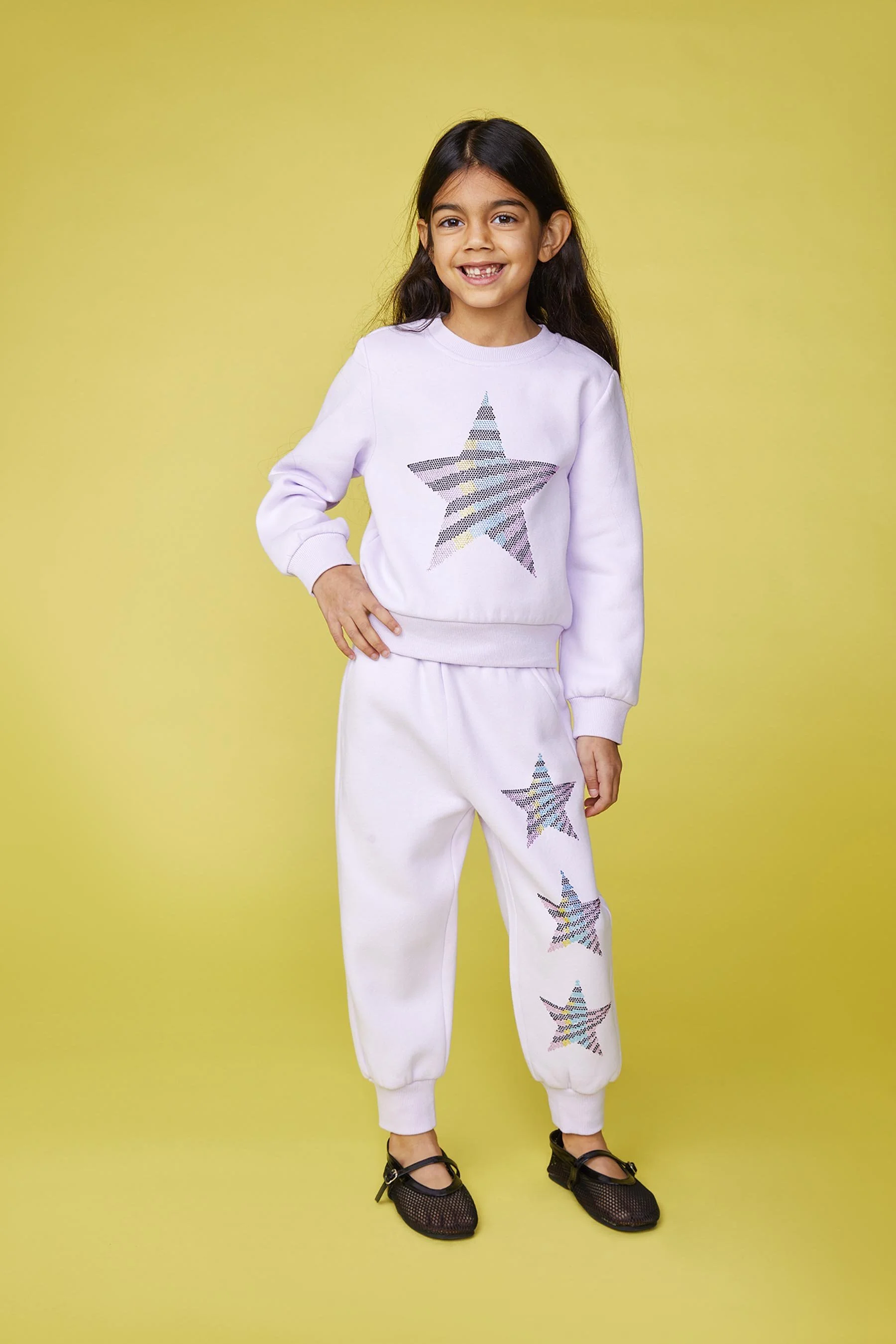 A young girl smiling while standing against a yellow background, wearing a white sweatshirt and sweatpants with multicolored star patterns, and black shoes.