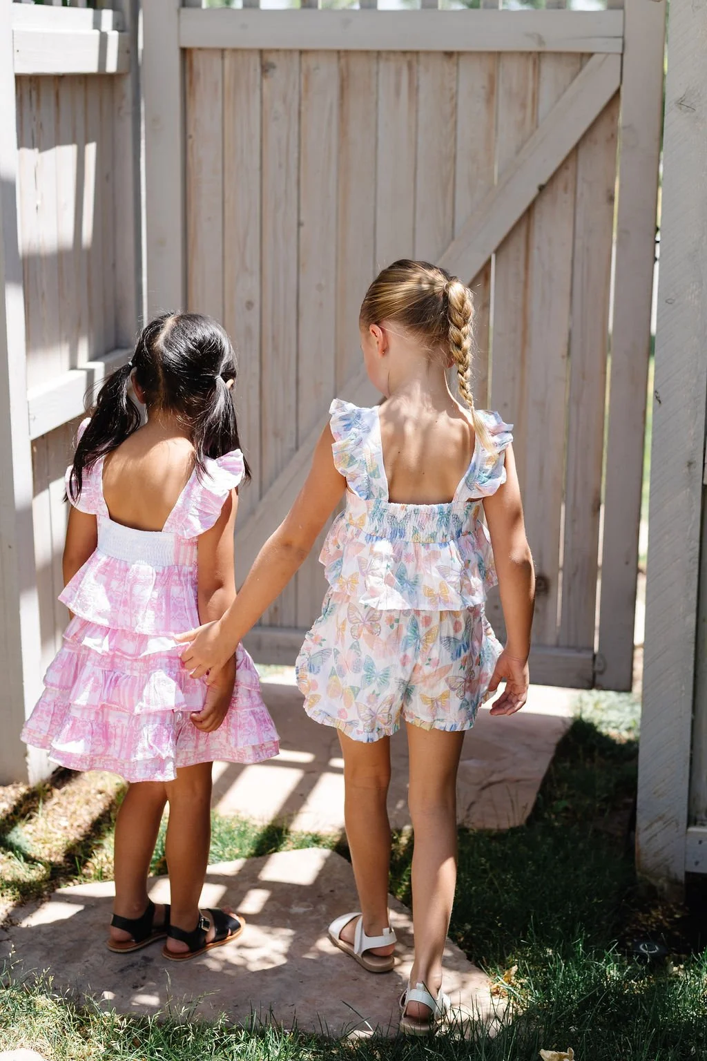 Two young girls holding hands walking through a wooden gate, wearing light-colored summer dresses with ruffles.