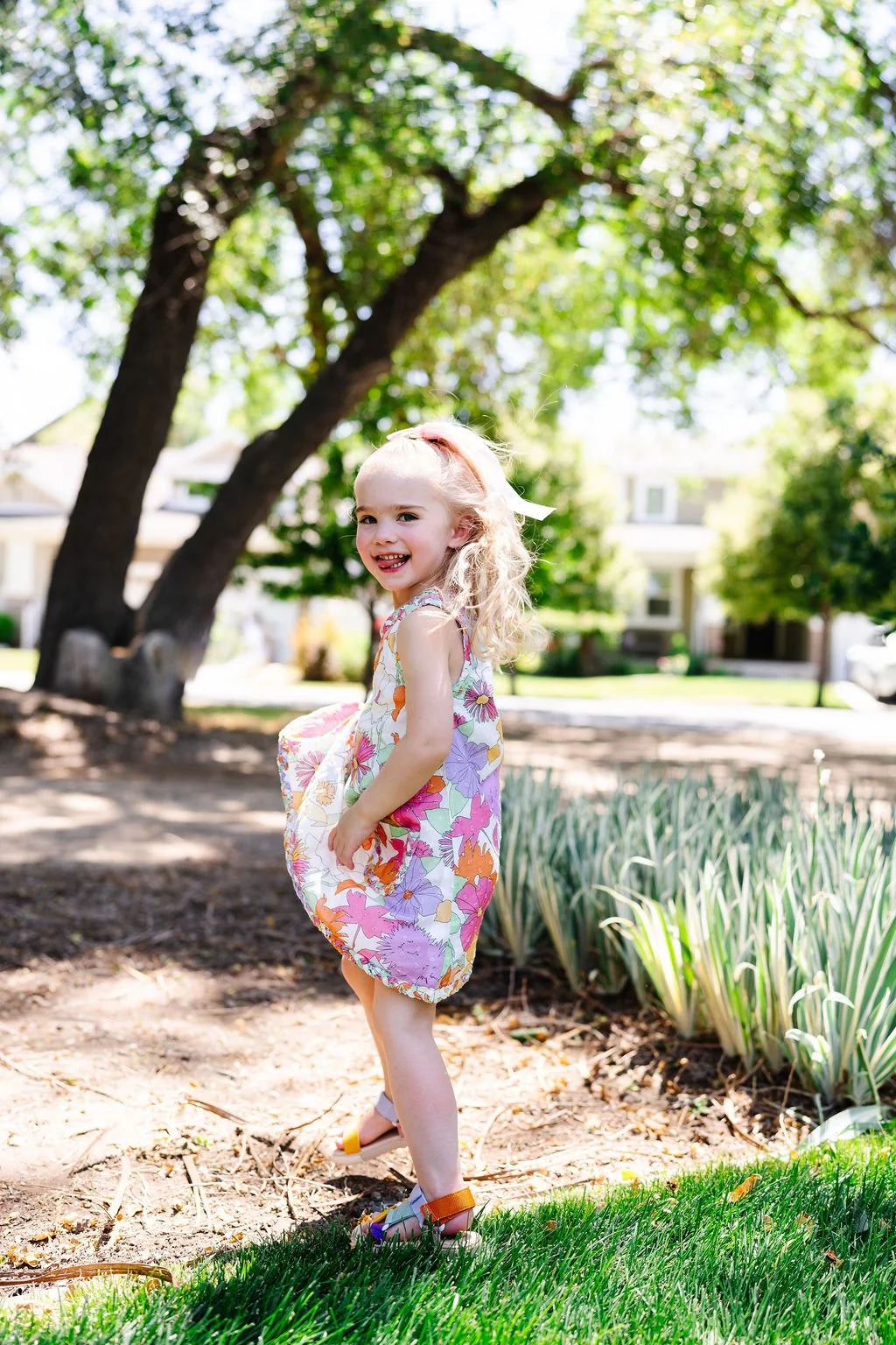 A young girl with curly blonde hair and a floral dress is standing outdoors in a sunny garden or park, smiling and holding her dress, with large trees and a house in the background.