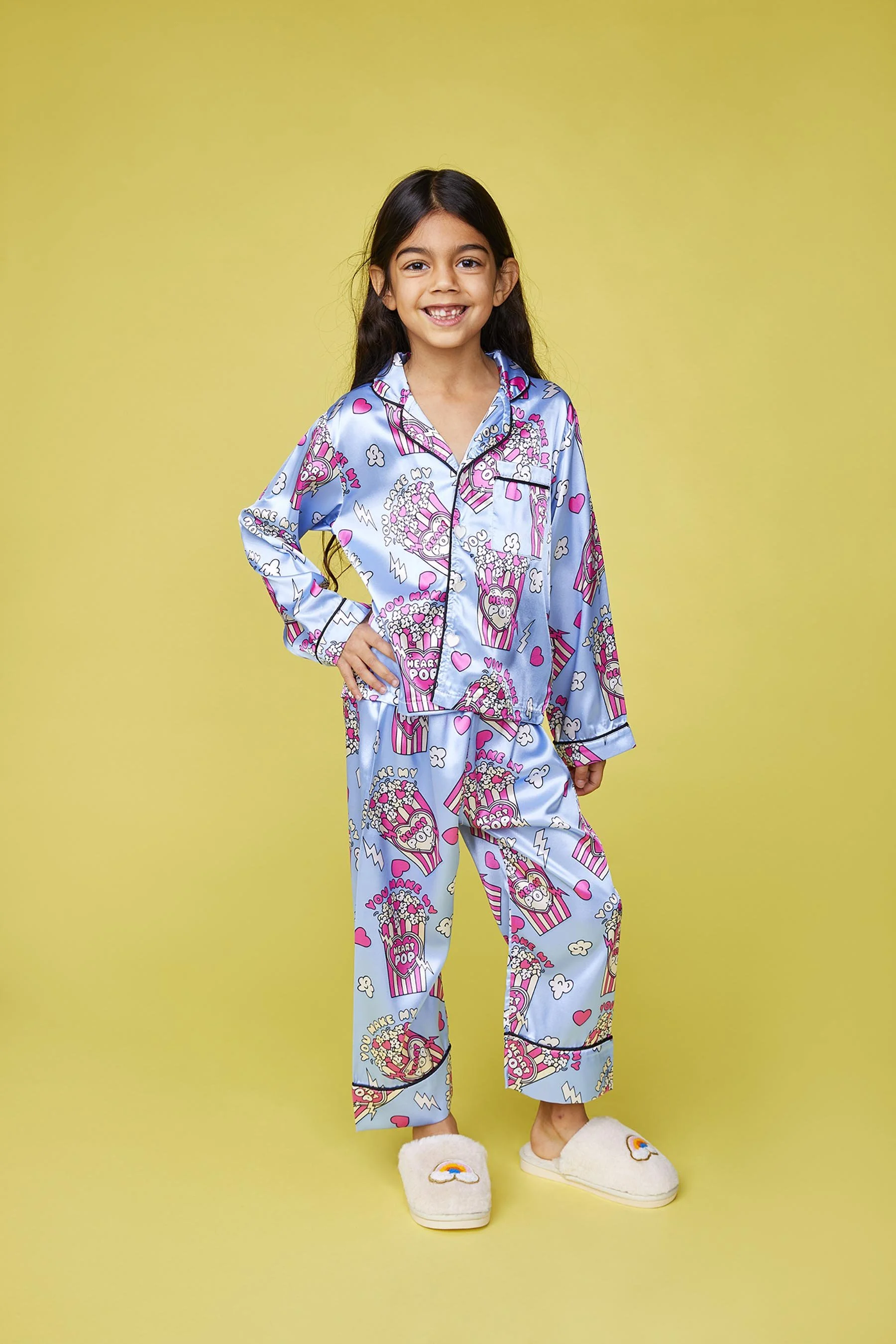 A young girl standing against a yellow background, wearing blue pajamas with popcorn, hearts, and popcorn illustrations, and cream-colored slippers with rainbow designs.
