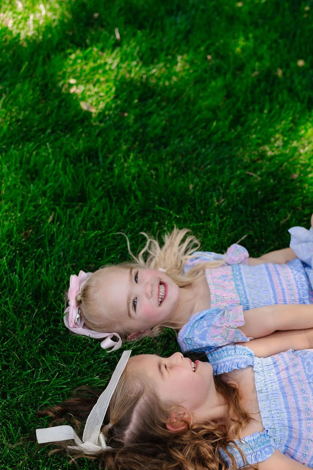 Two young girls lying on green grass, smiling and looking at each other.