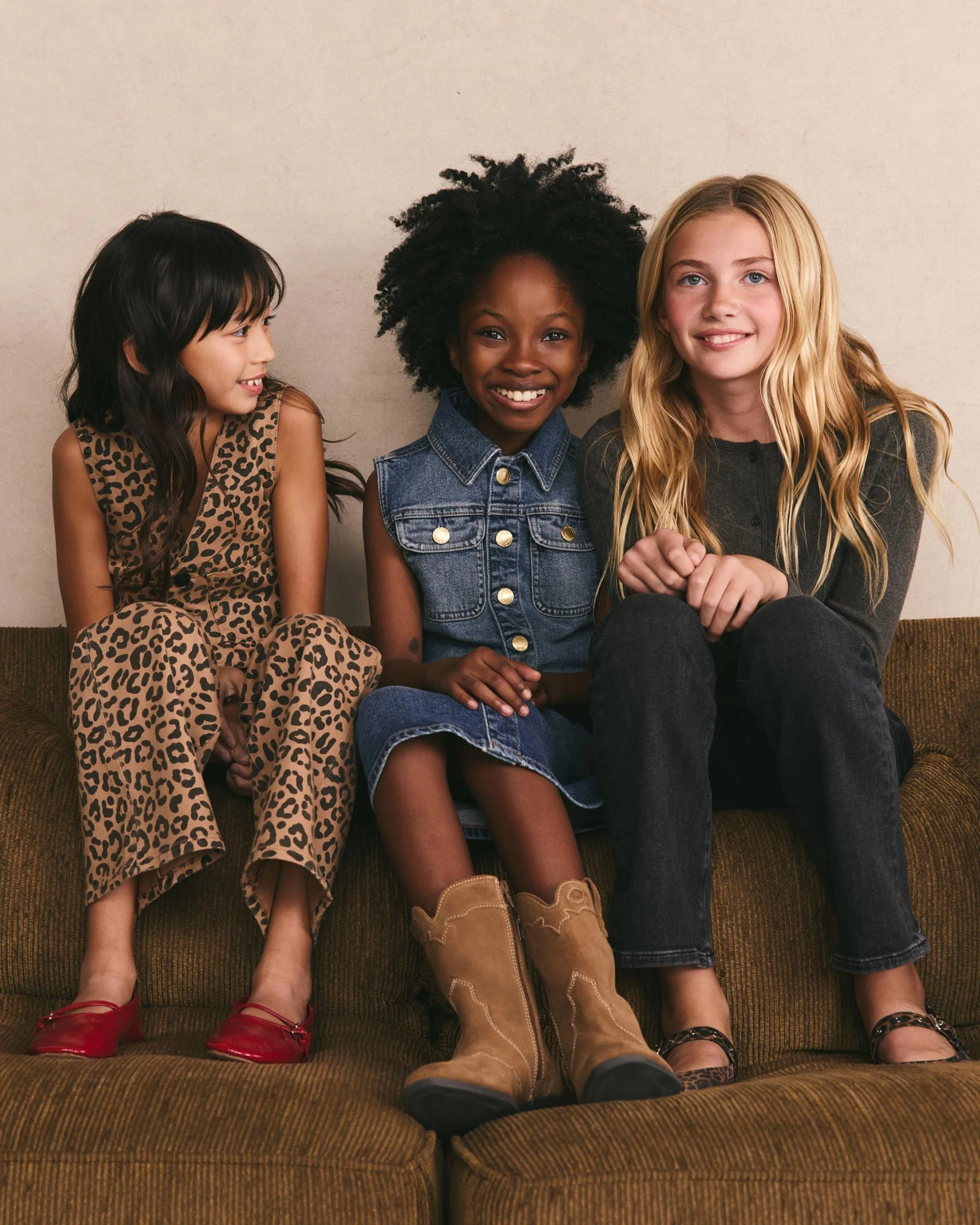 Three young girls sitting together on a brown sofa, smiling, dressed in casual outfits, with a plain beige wall in the background.