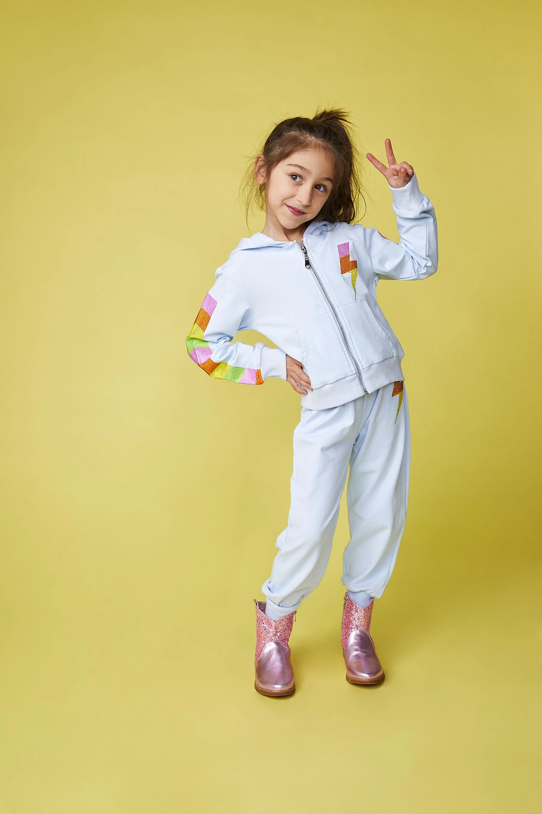 A young girl standing against a yellow background, wearing a white tracksuit with colorful rainbow lightning bolt patches and pink glitter cowboy boots. She has her right hand on her hip and is making a peace sign with her left hand near her head.
