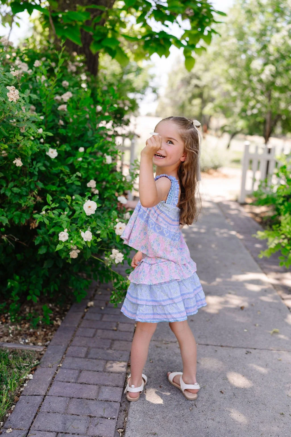 A young girl with long wavy brown hair, wearing a pastel-colored ruffled dress and white sandals, is smiling and playing with a flower petal in a garden next to a blooming bush with white flowers. She is standing on a paved pathway in a shaded outdoo