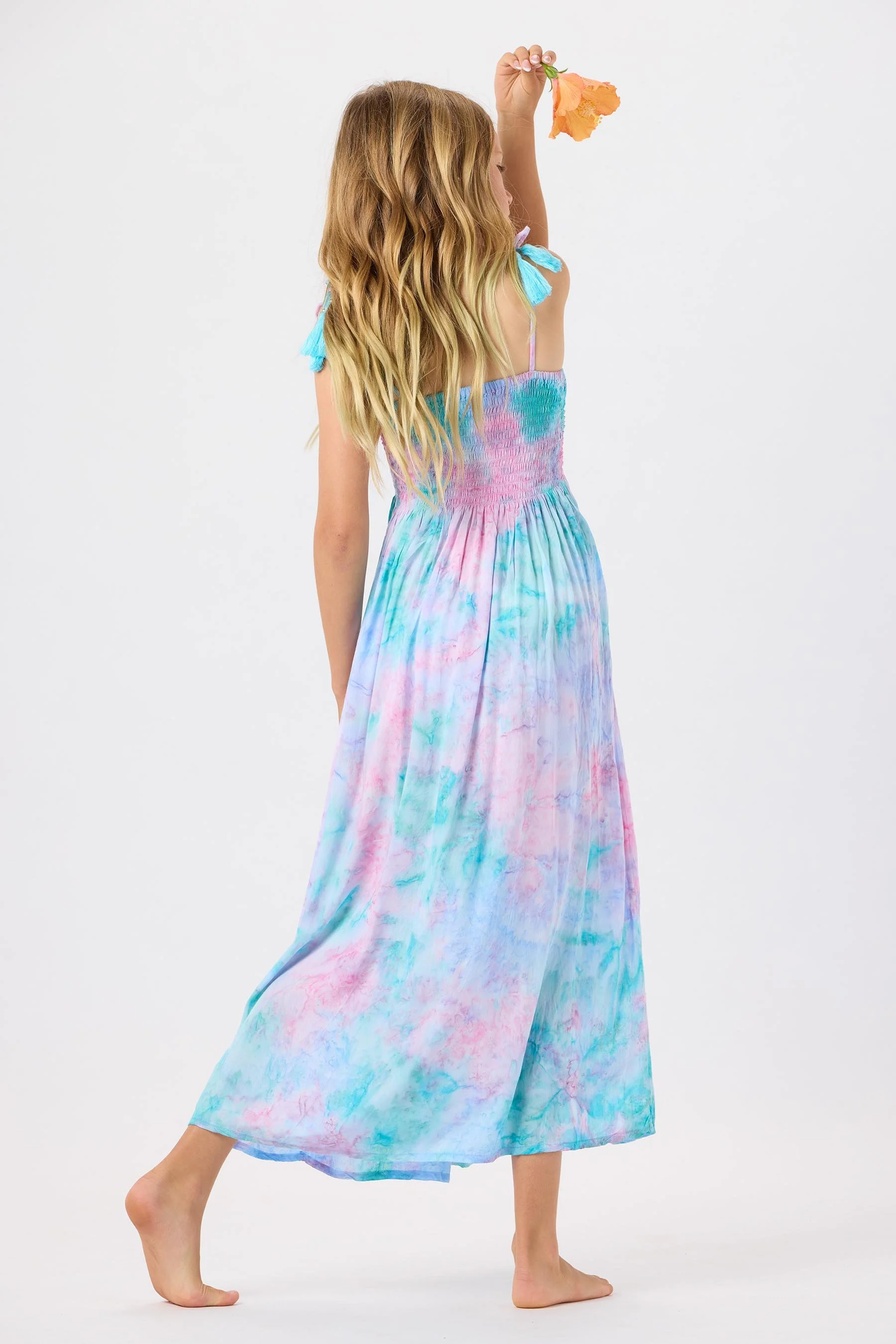 A young girl with long blonde hair is holding an orange flower up to her head. She is wearing a colorful, tie-dye maxi dress with spaghetti straps and blue tassel accents on the shoulders, standing barefoot against a plain white background.