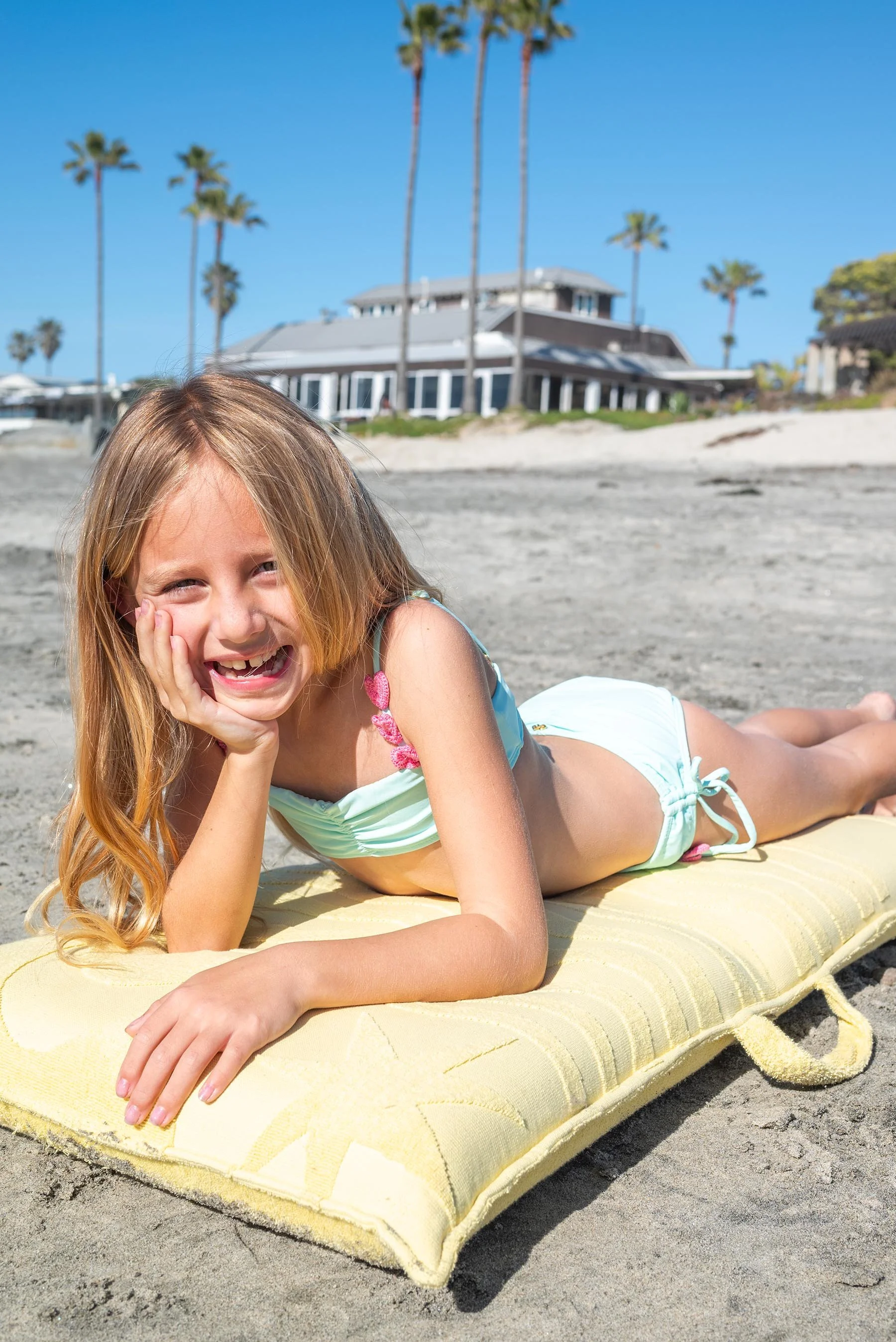 A young girl with red hair smiling and lying on a yellow towel on a sandy beach, wearing a turquoise swimsuit with pink accents, with a large house and palm trees in the background under a clear blue sky.
