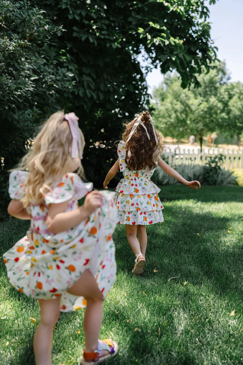 Two young girls wearing matching fruit-print dresses playing outdoors in a grassy yard with trees and a white picket fence in the background.
