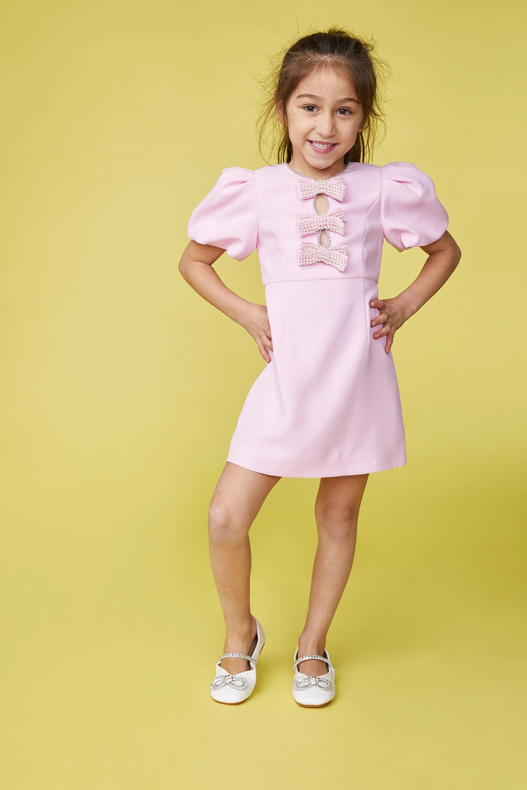 A young girl wearing a pink dress with puffed sleeves and three decorative bows down the front, standing against a yellow background.