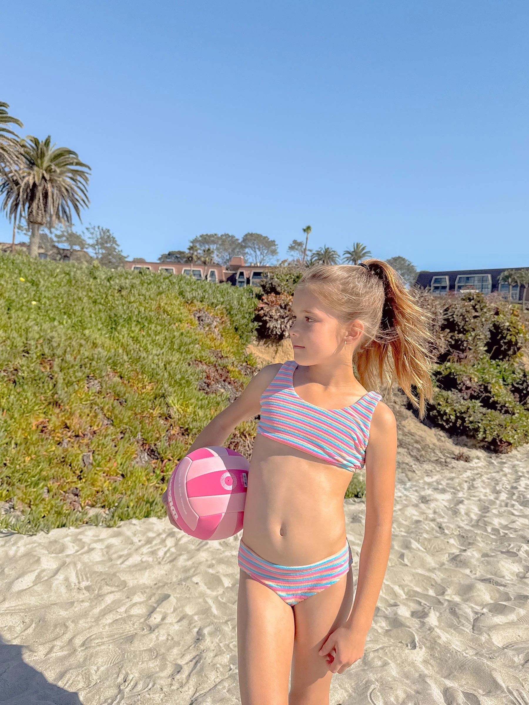 A young girl stands on the beach holding a pink and white beach ball, wearing a striped swimsuit, with palm trees and buildings in the background.