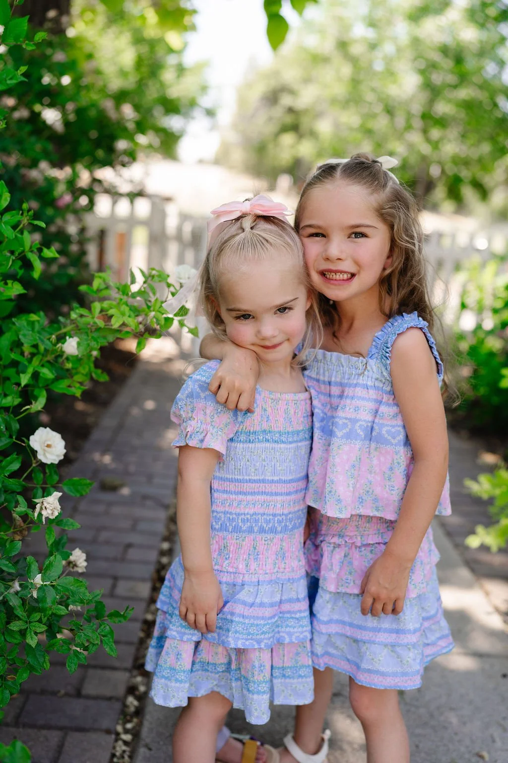 Two young girls hugging and smiling outdoors on a garden pathway surrounded by green bushes with white flowers.