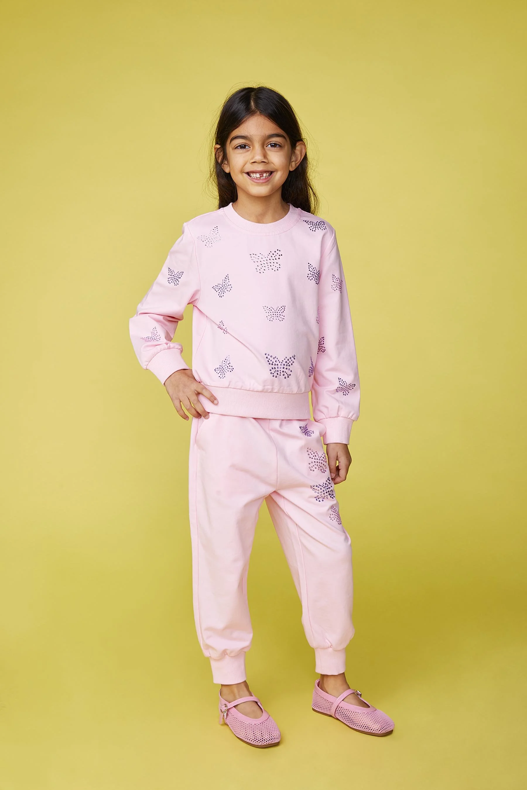 A young girl with dark hair in a ponytail, smiling, wearing a pink sweatshirt and matching pink sweatpants with purple butterfly patterns, standing against a yellow background.