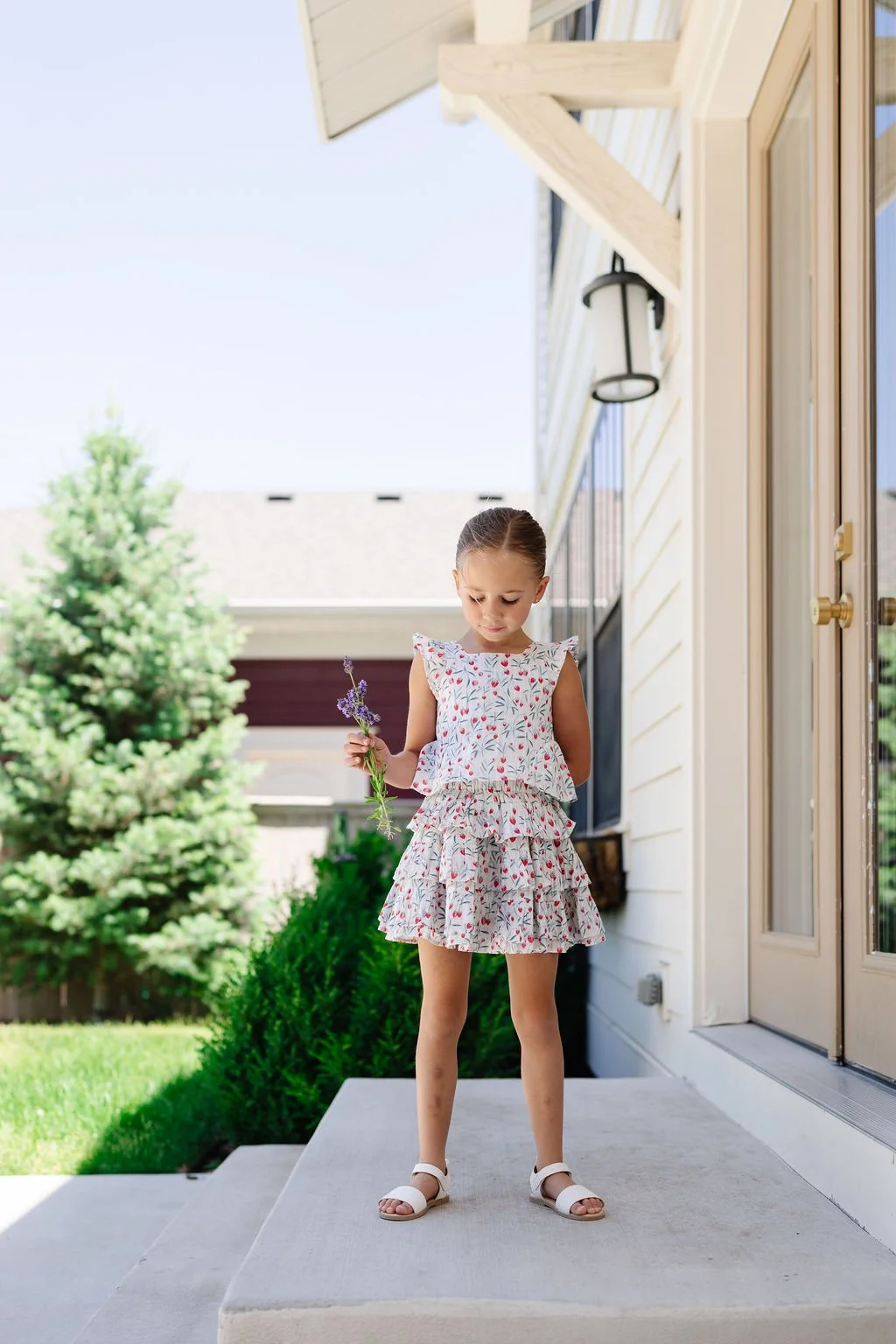 A young girl wearing a floral dress and white sandals standing on a porch holding a small bunch of purple flowers, with a suburban home and green bushes in the background.