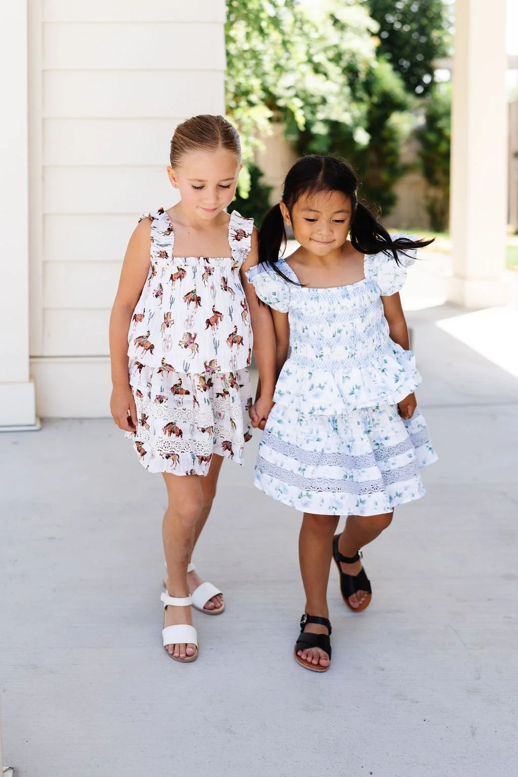 Two young girls holding hands and walking outdoors on a sunny day, wearing dresses with floral and animal patterns.