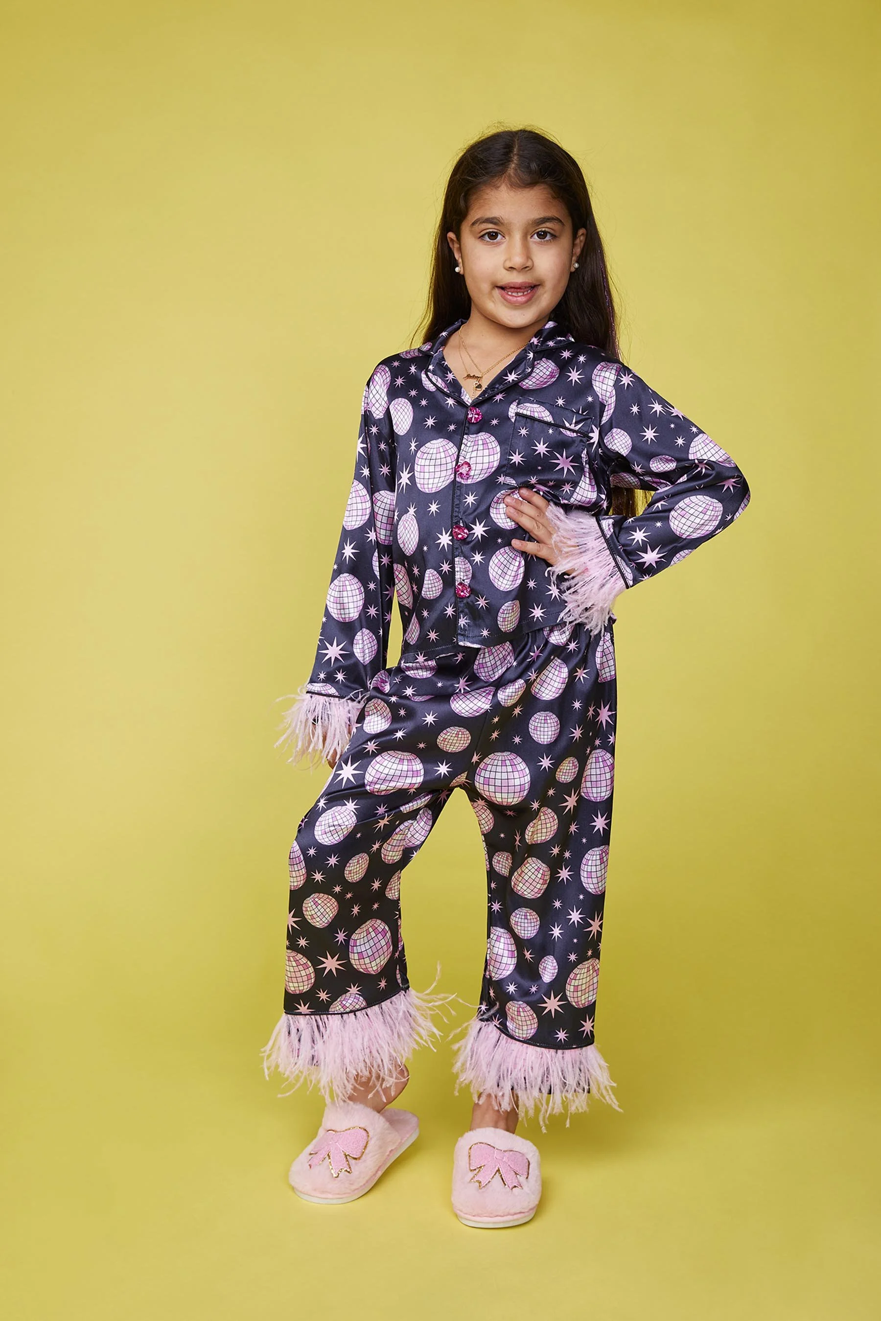 Young girl in navy pajama set with pink and white geometric patterns, pink feather trim, and pink slippers standing against a yellow background.